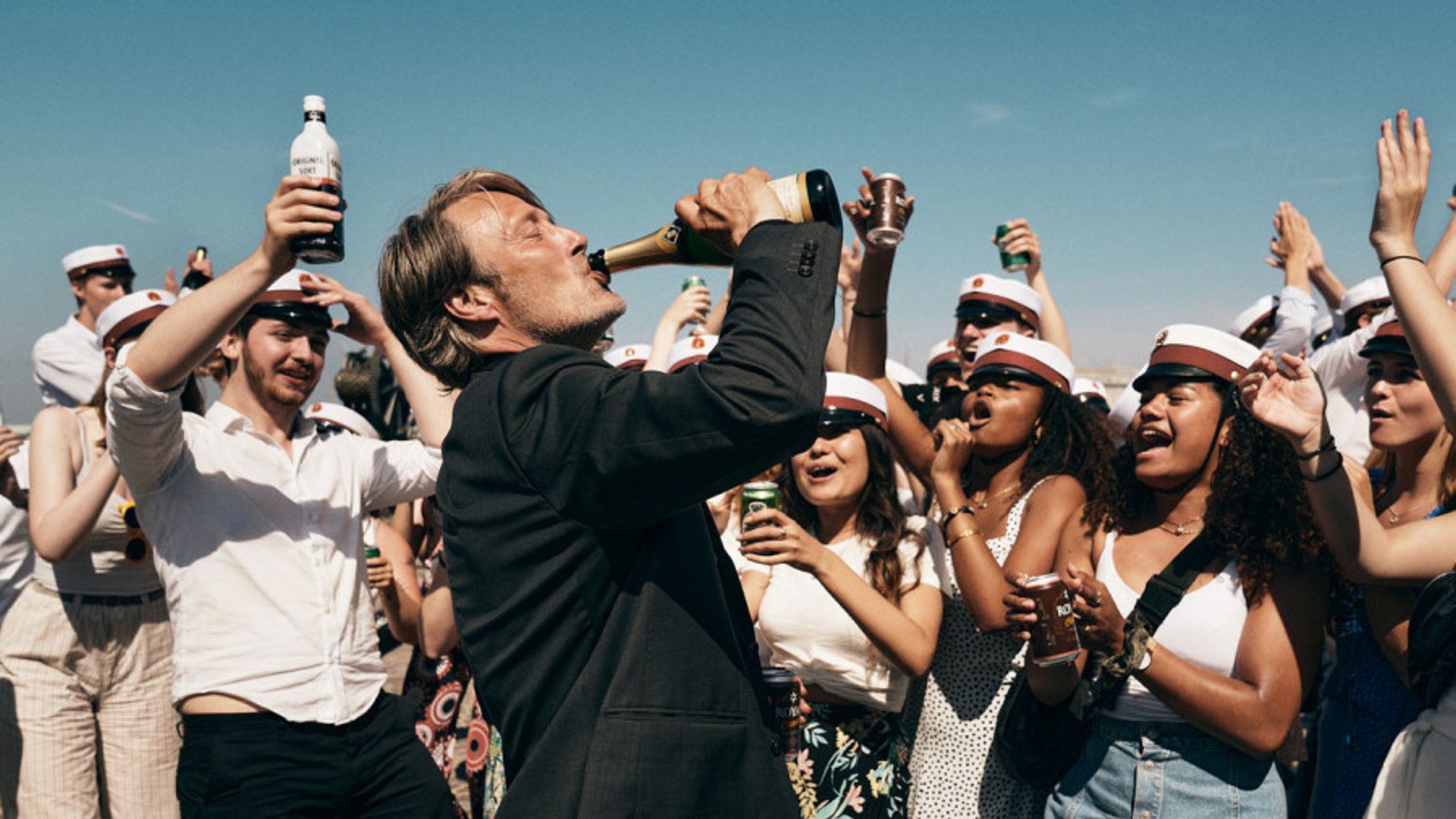 a crowd cheers on mads mikkelsen as he drinks from a bottle