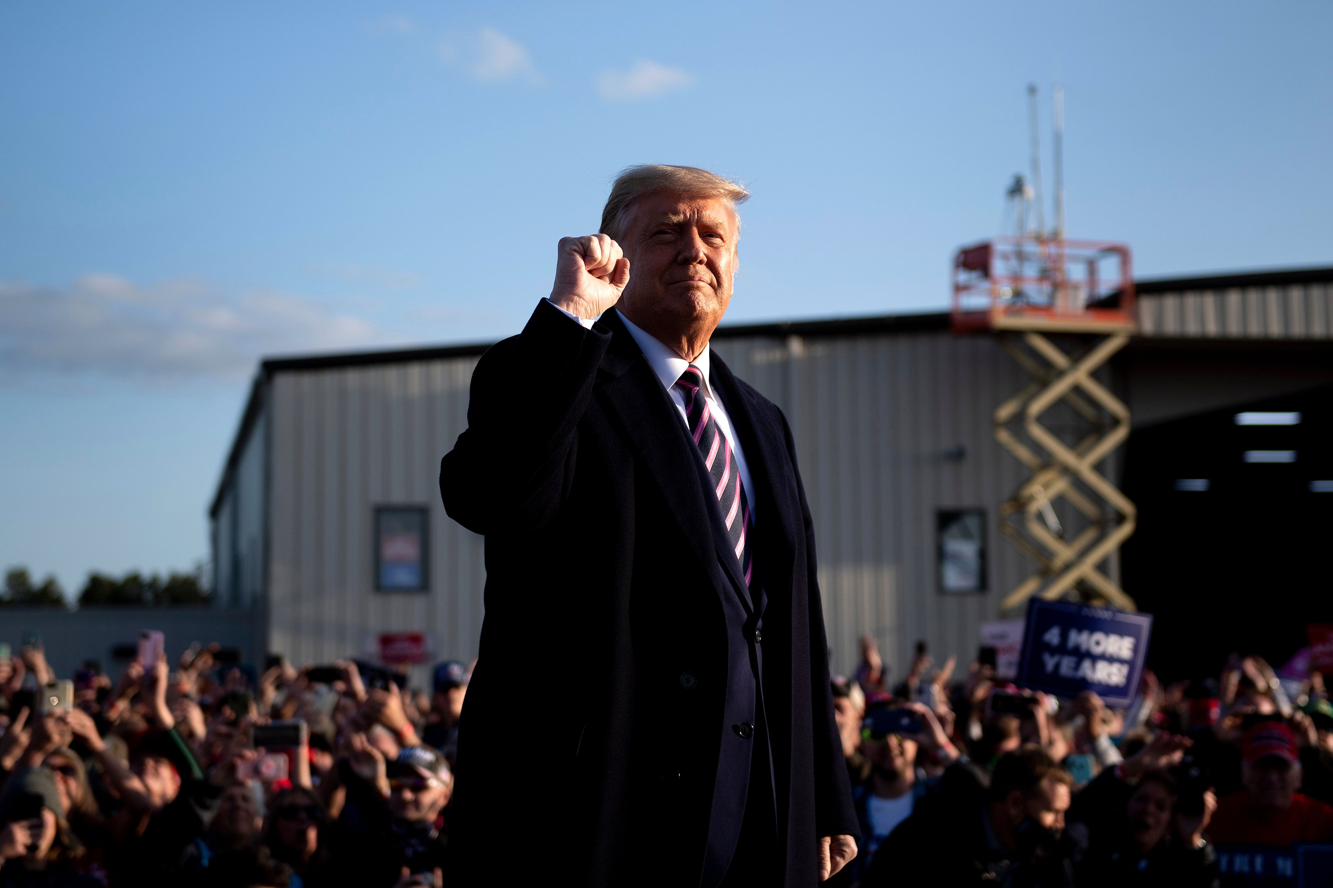 Trump, in a red, white, and blue striped tie and back winter coat, smiles slightly, lit by the setting sun. He raises his right fist; behind him, a large crowd cheers, many holding Trump 2020 signs.