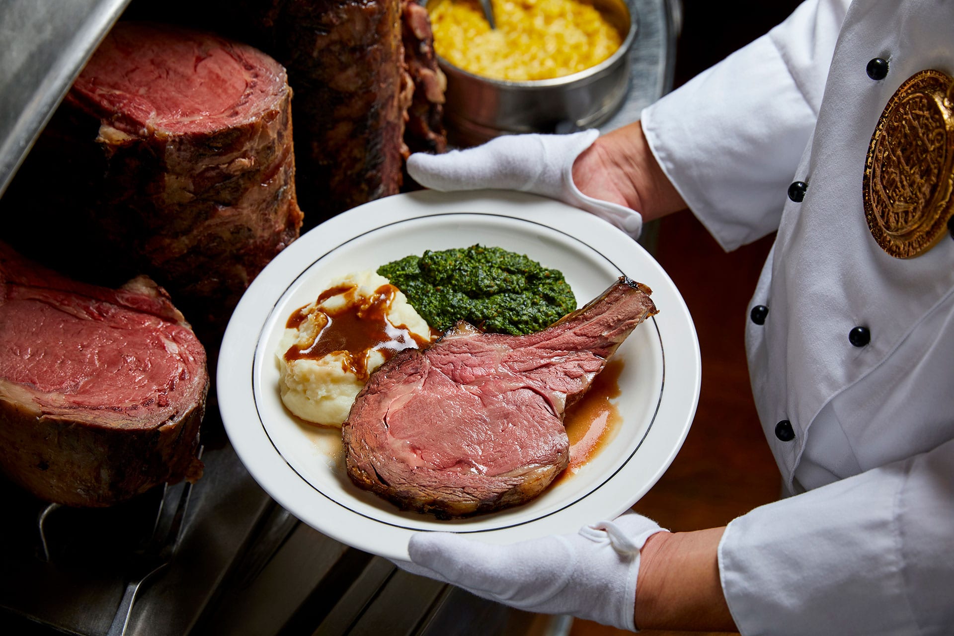 A chef in a white jacket and white gloves holds a plate of sliced prime rib, mashed potatoes, and green vegetables.