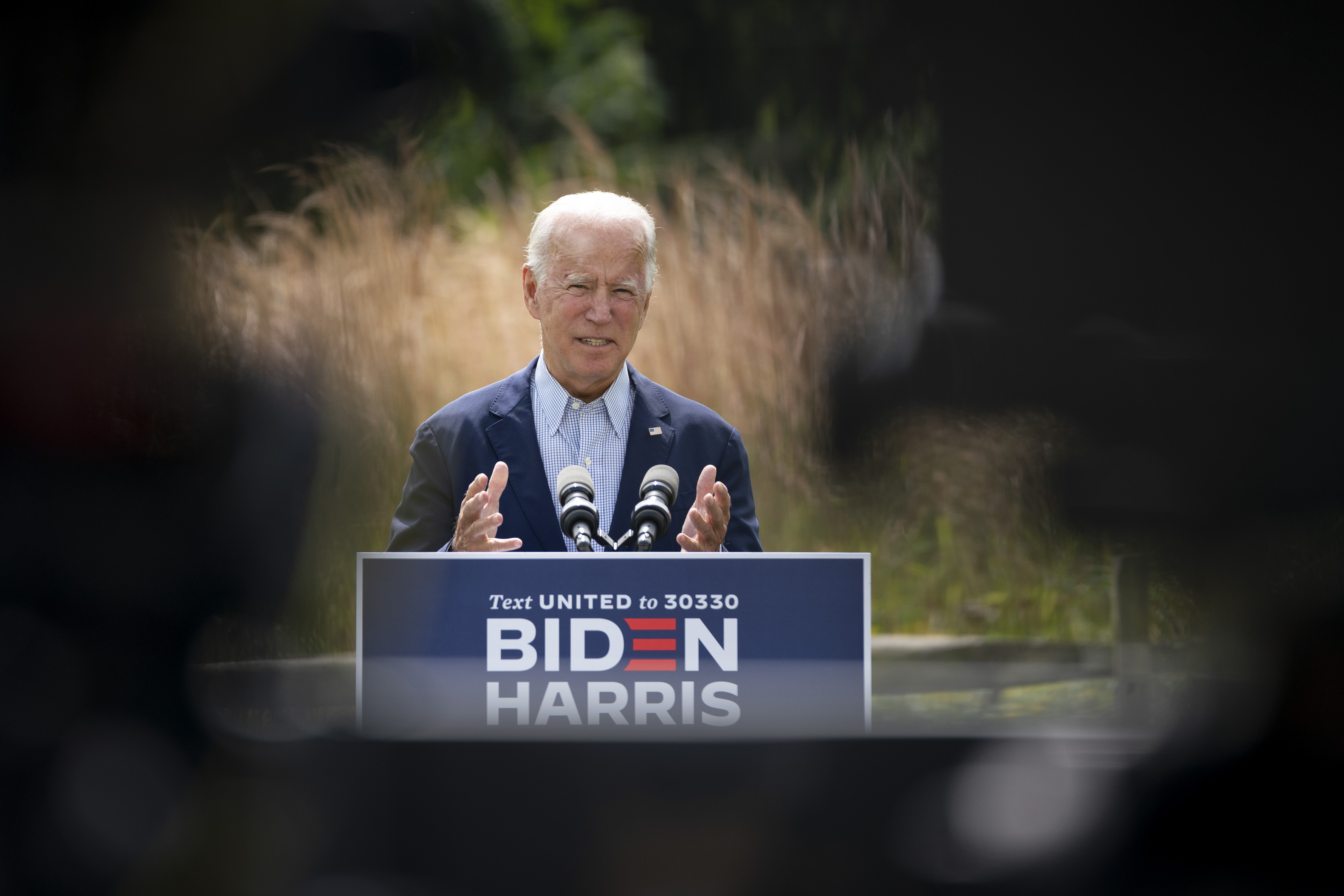 Man wearing suit stands behind podium reading “Biden/Harris” in a field.