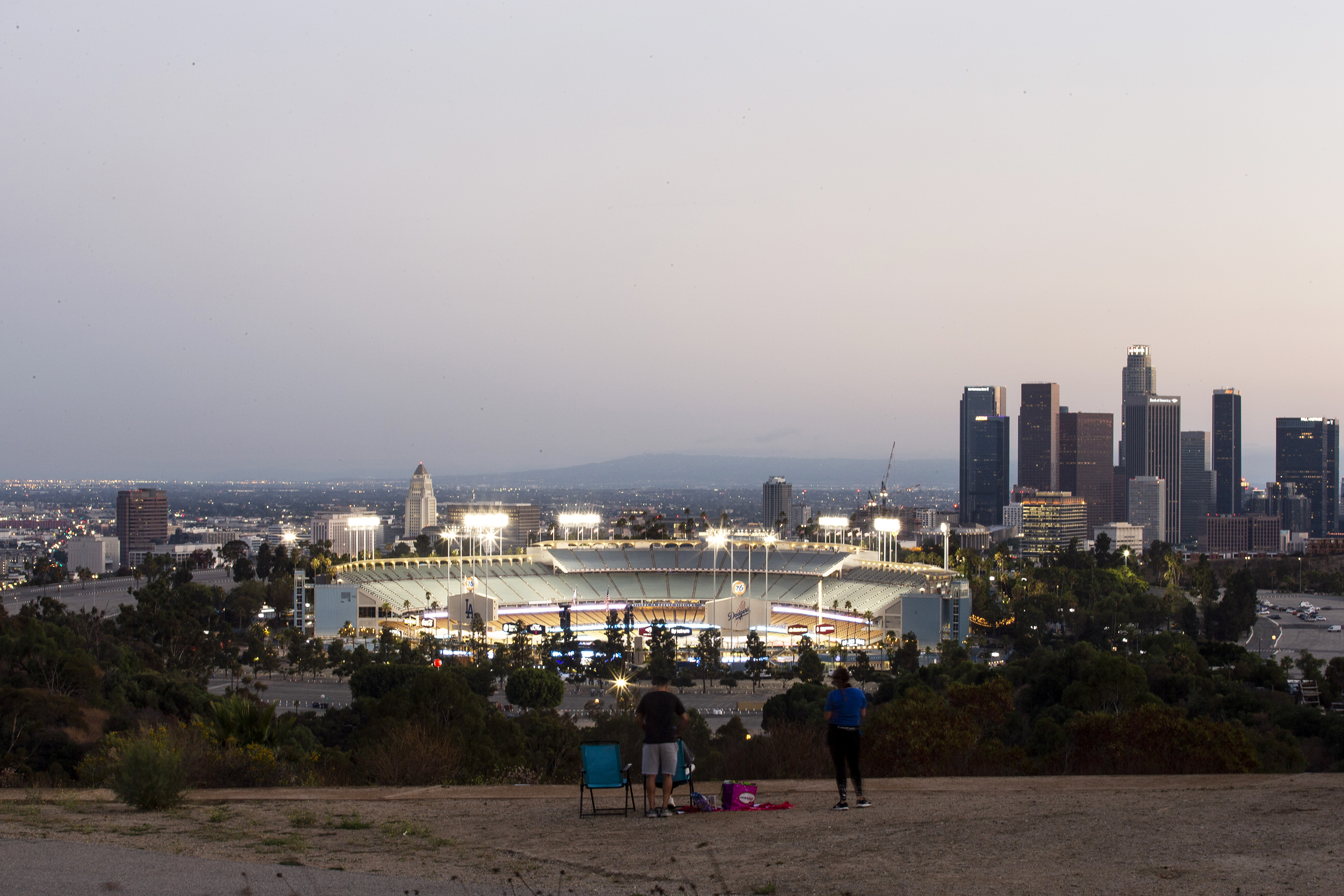 A family sits on chairs in a parking lot above the lights of Dodger Stadium, where teams are playing but no fans are in the stand, with the skyline of downtown LA behind.