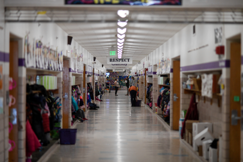An elementary school hallway with jackets on hooks and lunch bags on shelves, with two adults in the distance. Banners, one saying “Respect,” hang from the ceiling between lights.