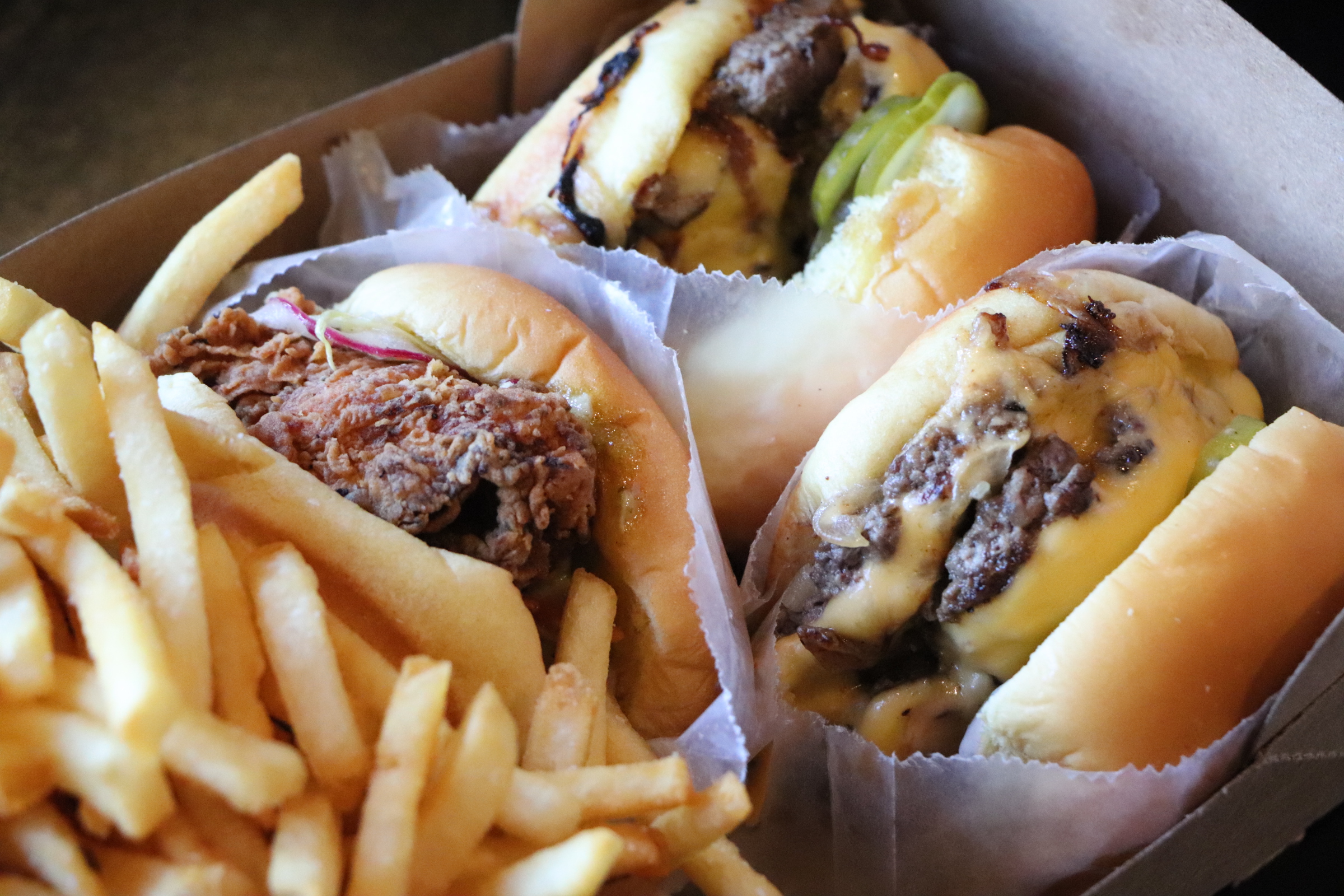 A close-up shot of two burgers, fries, and a fried chicken sandwich all in a paper bucket.