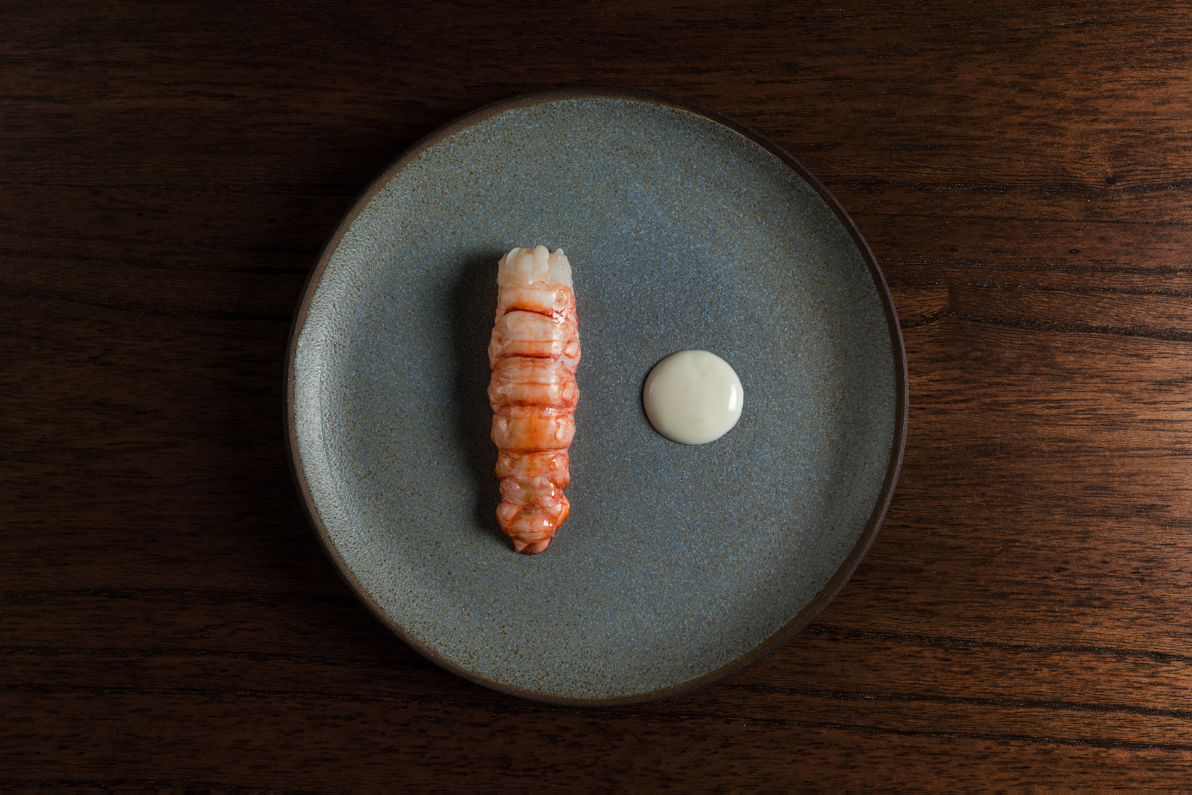 A piece of prawn or similar meat next to a small circle of white sauce on a gray stoneware plate on a wooden surface