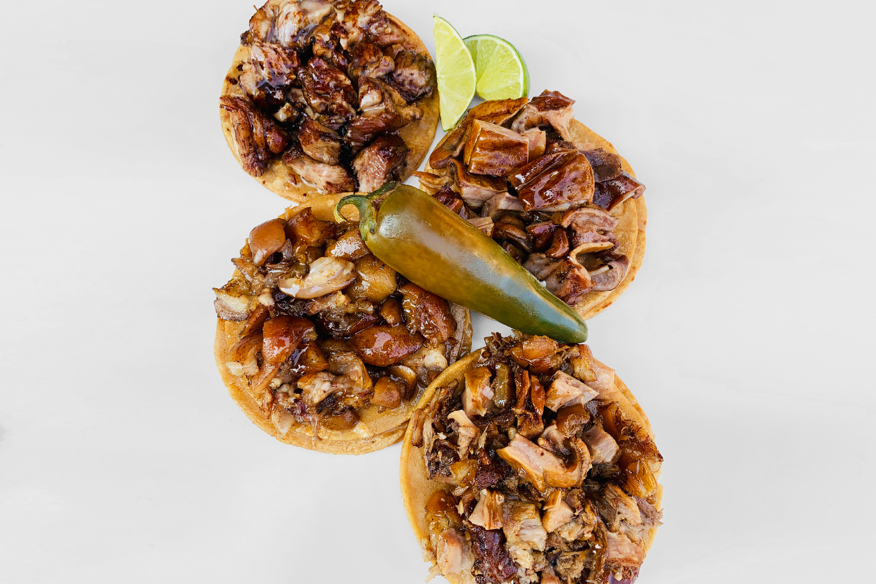 Four meat-laden tacos laid out on a white background with a large jalapeno pepper placed in the center