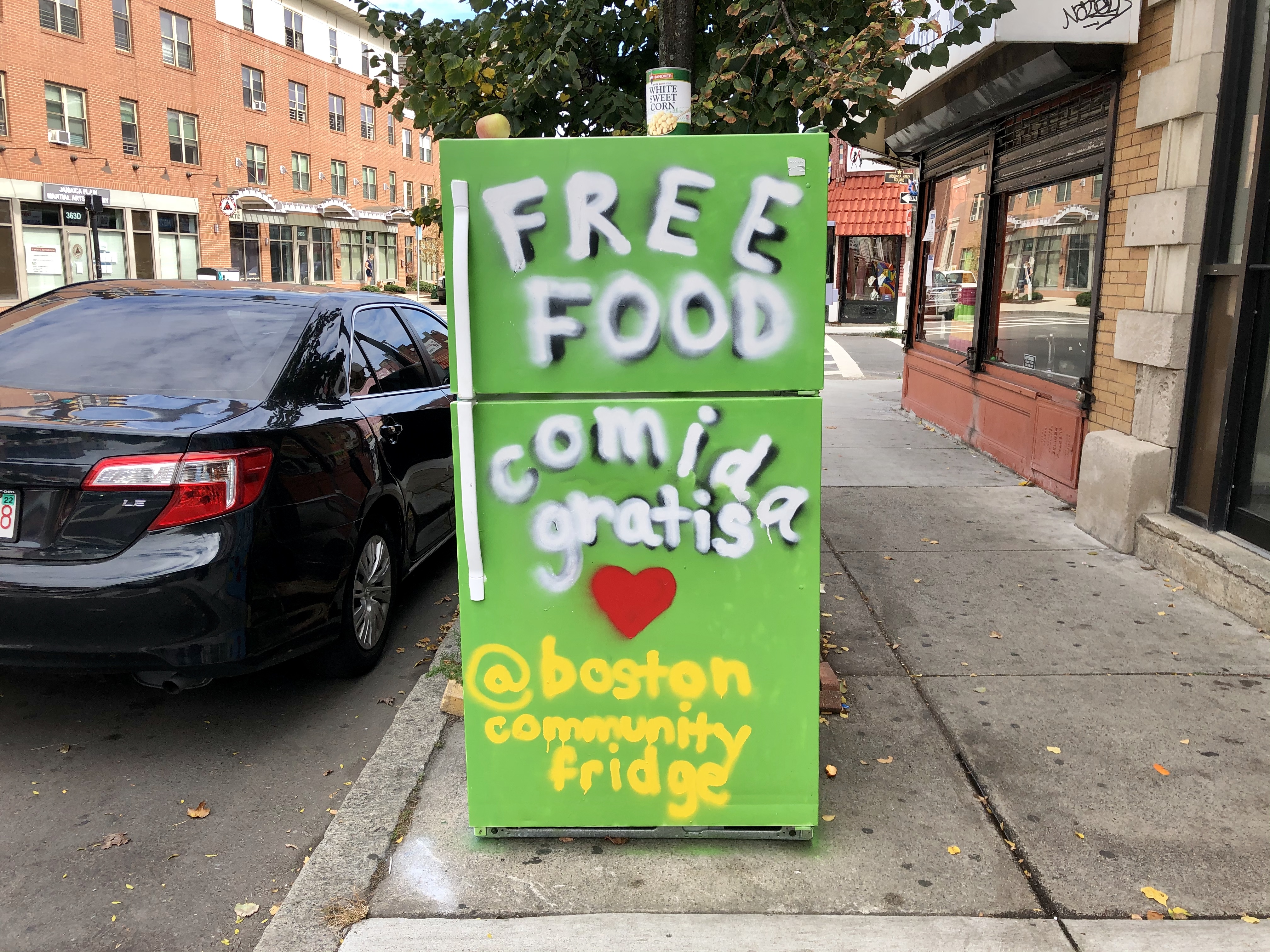 A lime green fridge with the words “free food” and “comida gratis” painted on the front doors sits on a sidewalk in Boston’s Jamaica Plain neighborhood.
