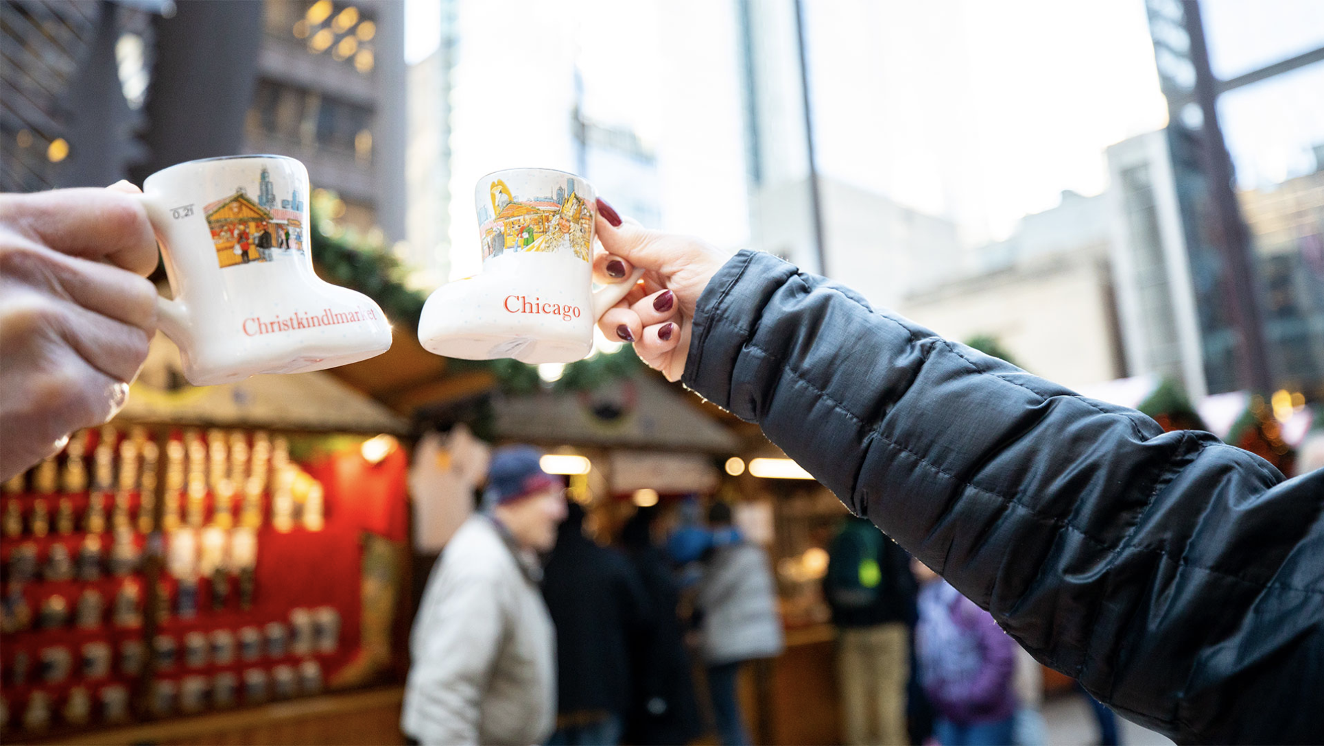 Two people holding small boot-shaped mugs cheers outside a Germanic-looking Christmas market stall.