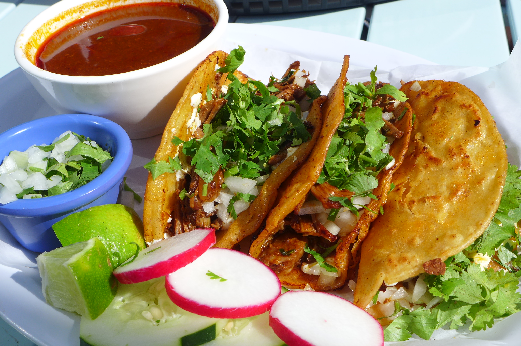 Three corn tortilla tacos with a dark soup on the side and garnished with bright red radish slices in foreground.