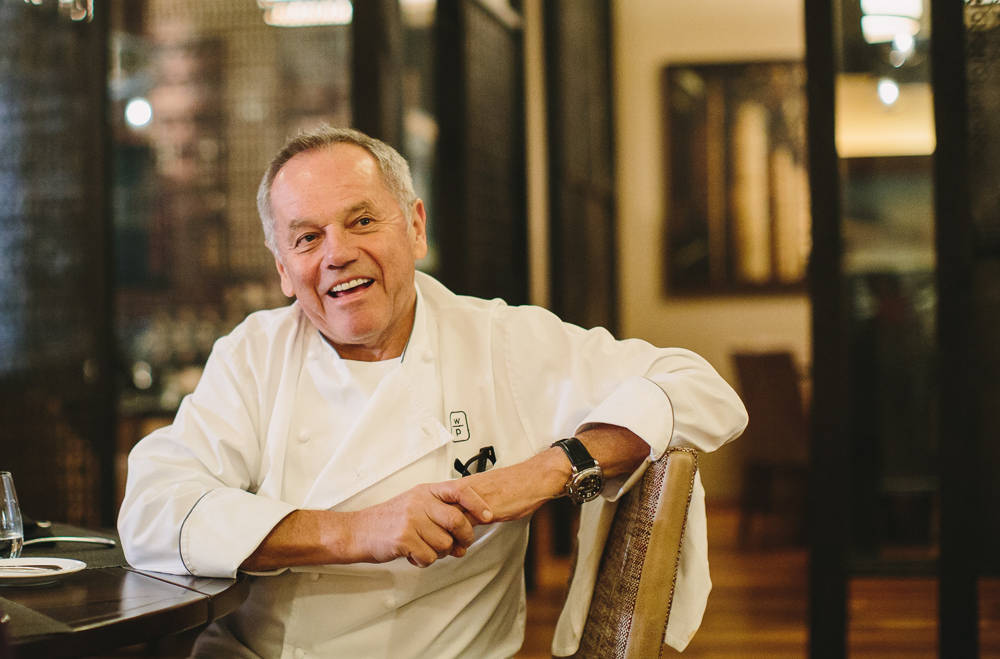 Wolfgang Puck sits in his chef jacket with his hands clasped and one arm leaning on the back of the chair.