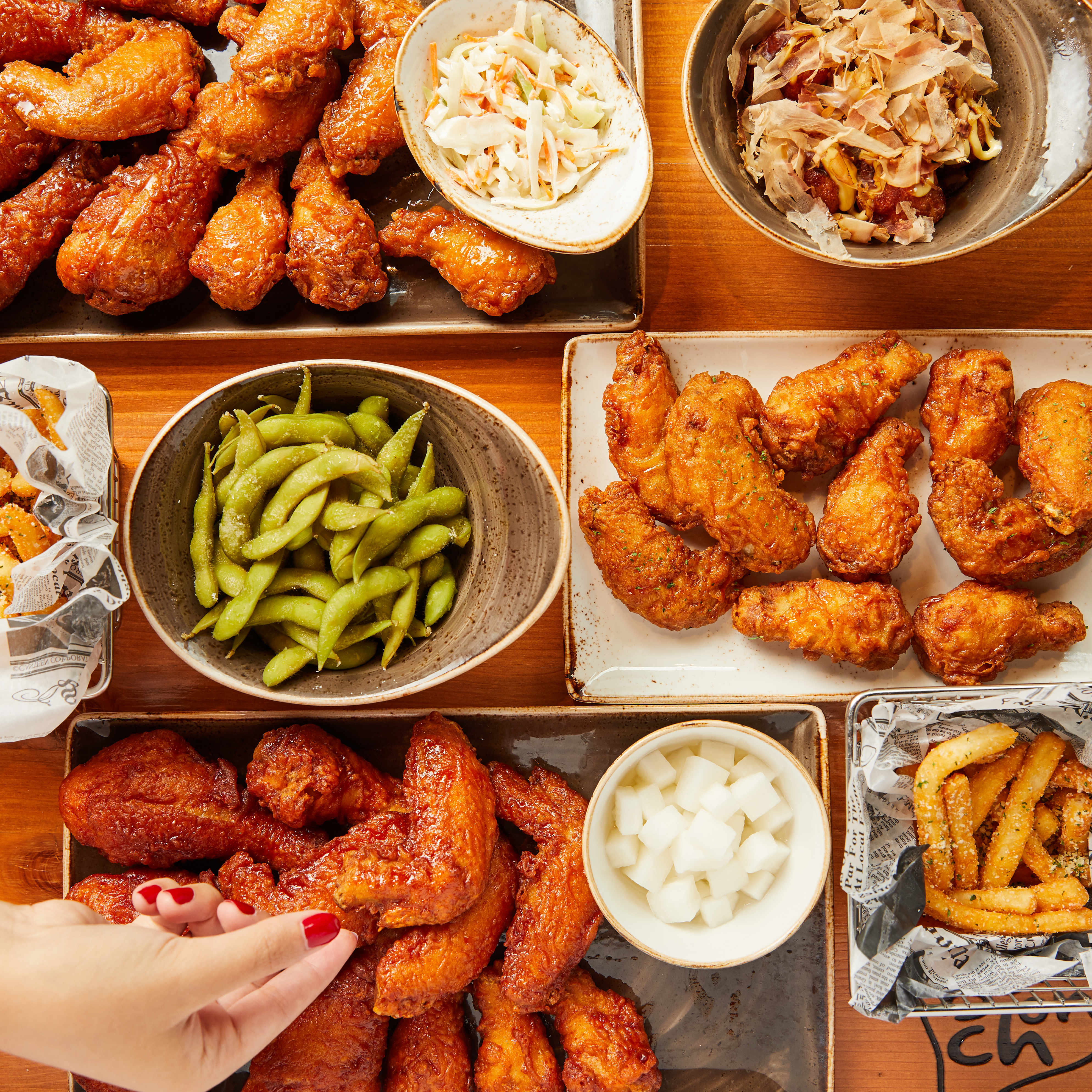 A wooden table is covered in plates of fried chicken, edamame, fries, and radish