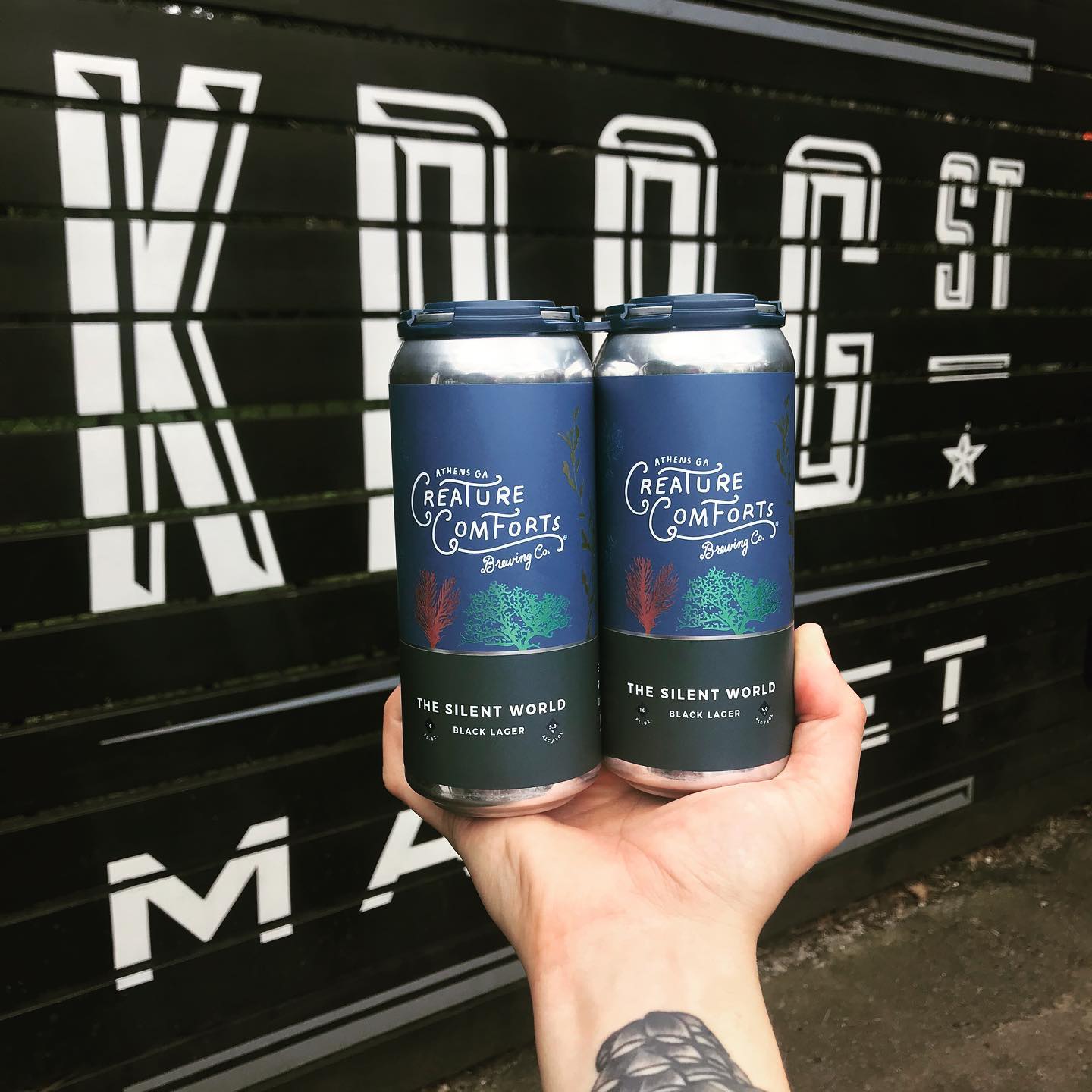 A hand with a tattooed arm holding two cans of Creature Comfort “The Silent World” black lager in front of the black painted wall with the white Krog Street Market logo on it