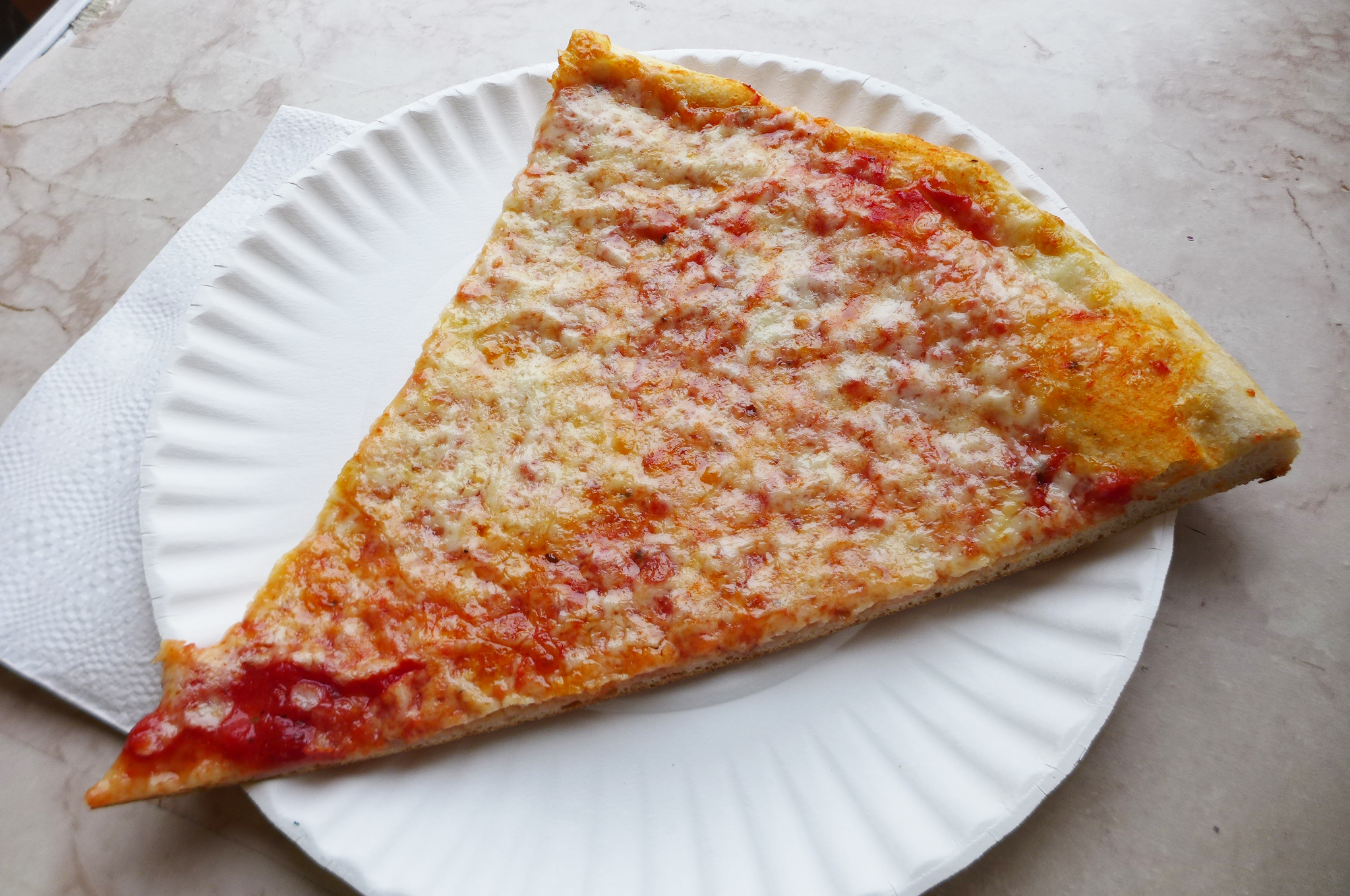A pizza slice with a modest amount of sauce and stippled with cheese on a paper plate.