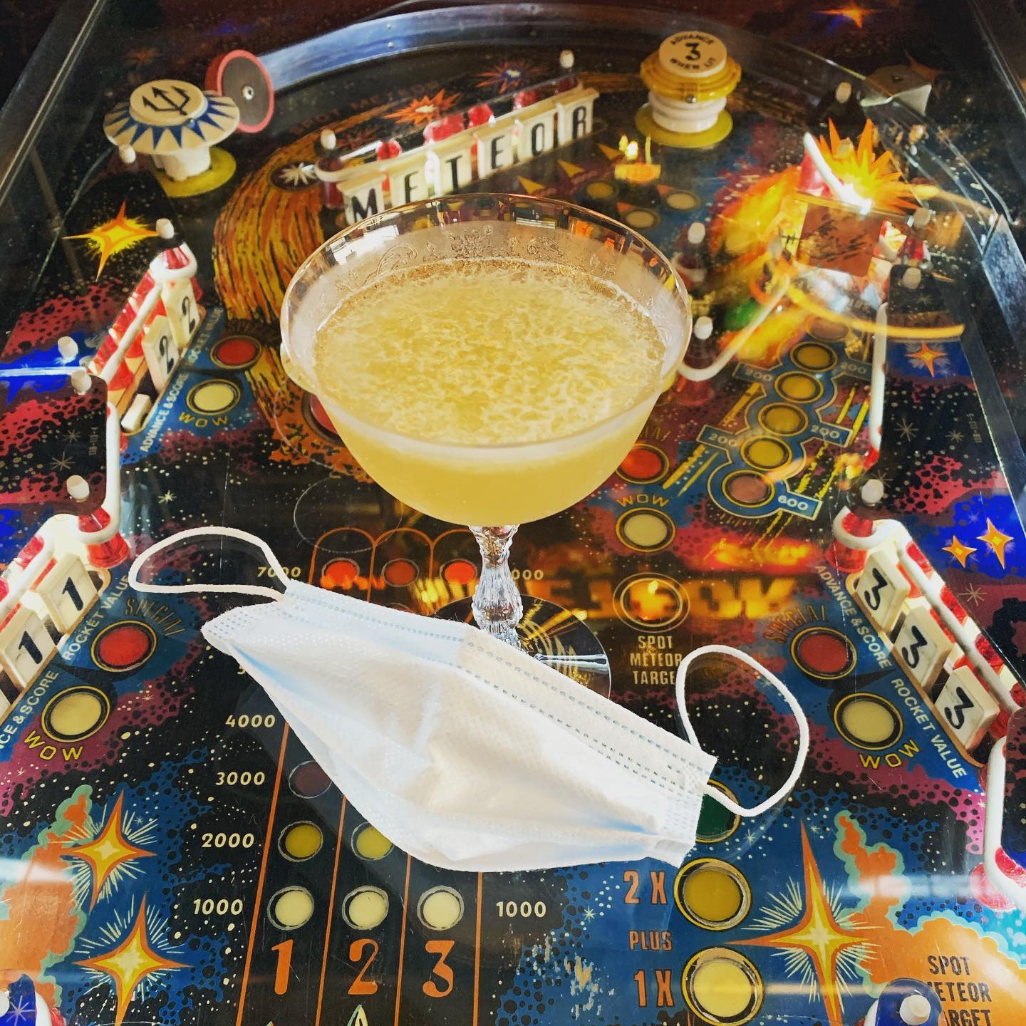 A martini glass with an icy yellow liquid sits on a vintage pinball machine with a blue, medical face mask in front of it