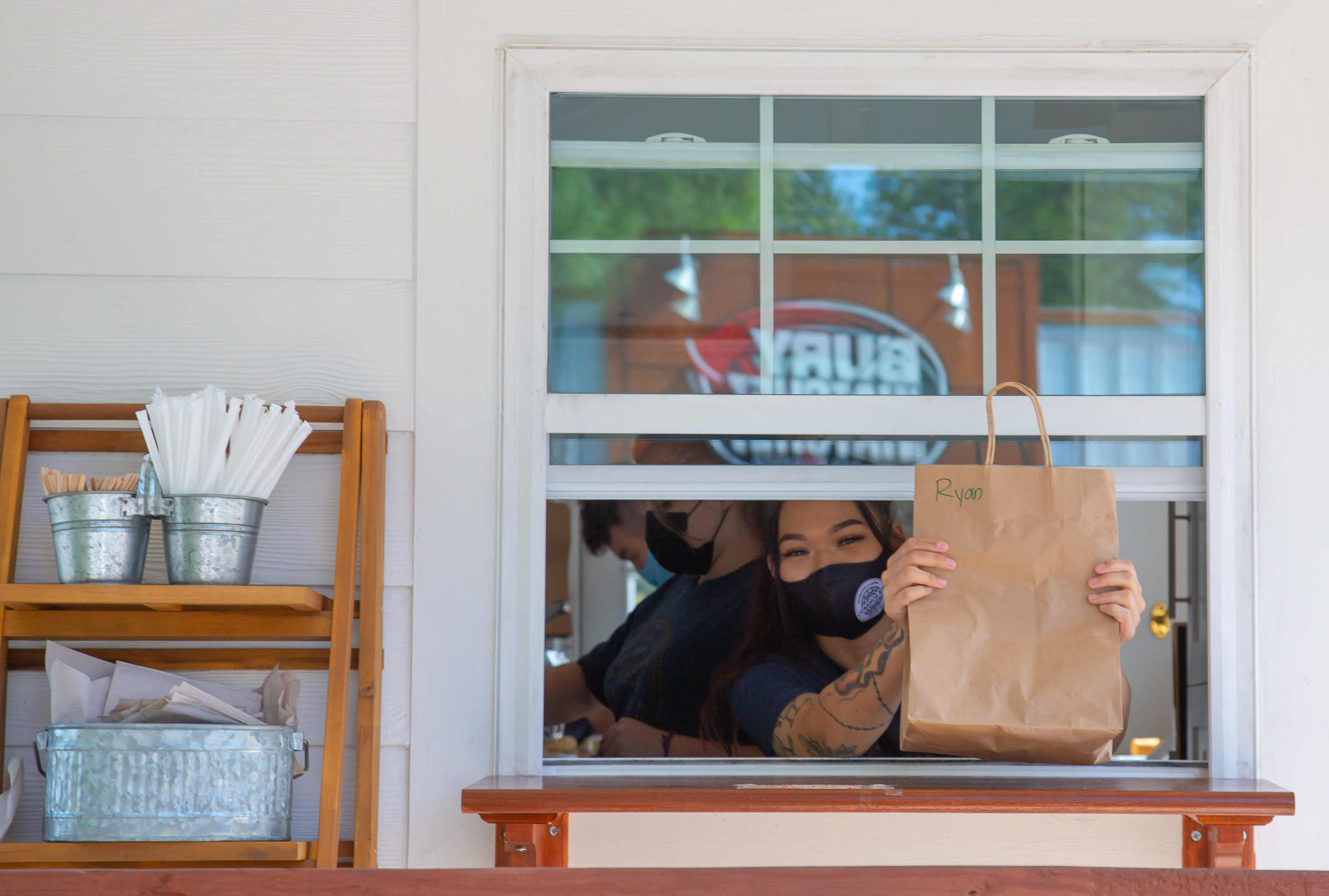 A woman of Asian descent in a black mask and tee shirt with tattooed arms handing a brown takeout bag through an open window. A wooden stand next to the window contains tins of straws, wooden coffee stirrers, and napkins