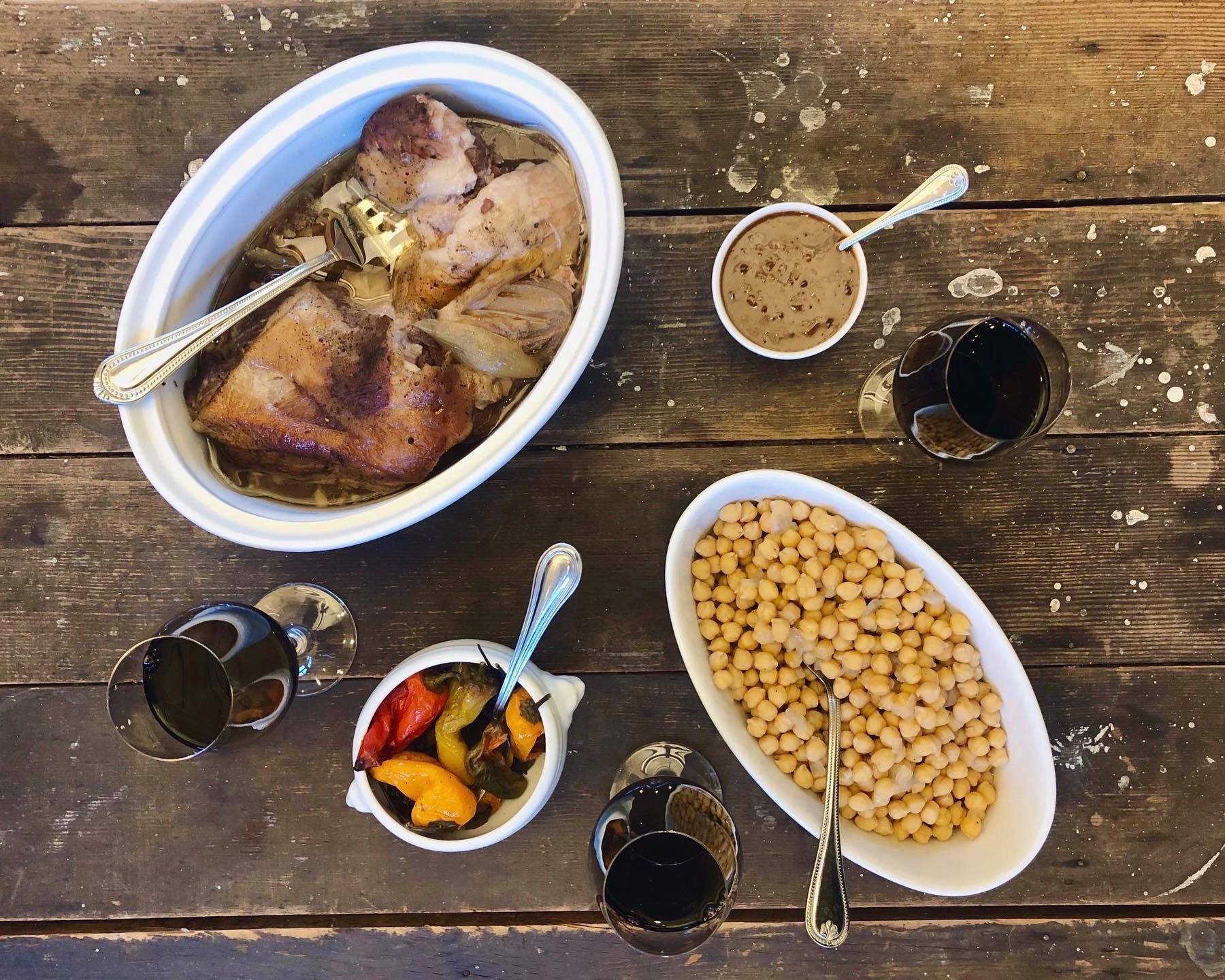A collection of dishes from Boat Street Kitchen, including a pot of roasted chicken, with three glasses of red wine on a rustic wood table