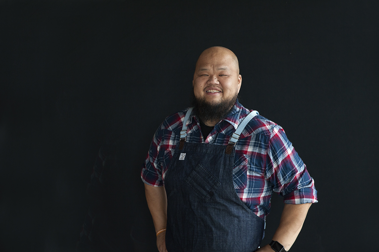 Chef Yia Vang is wearing a plaid shirt and denim apron, smiling, in front of a black background