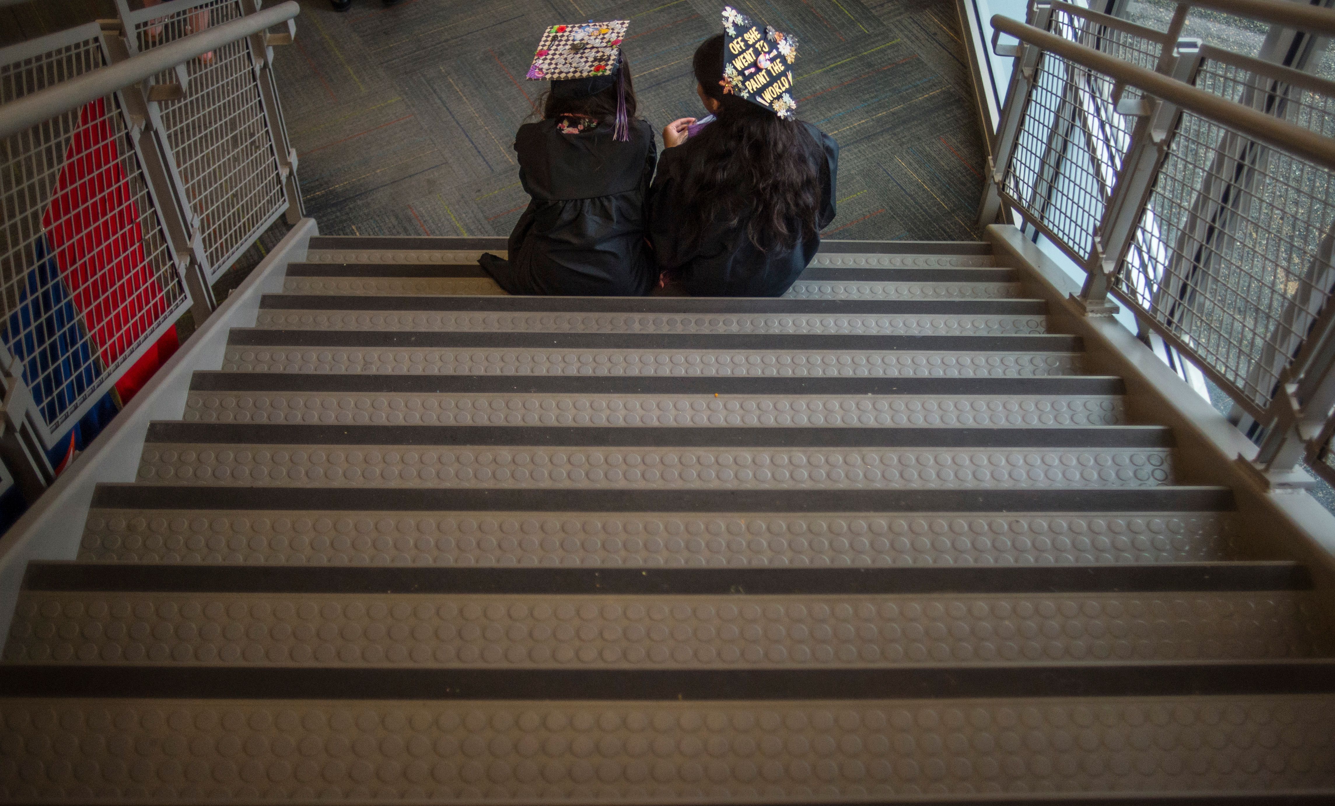 High school graduates in caps and gowns sit on a staircase.