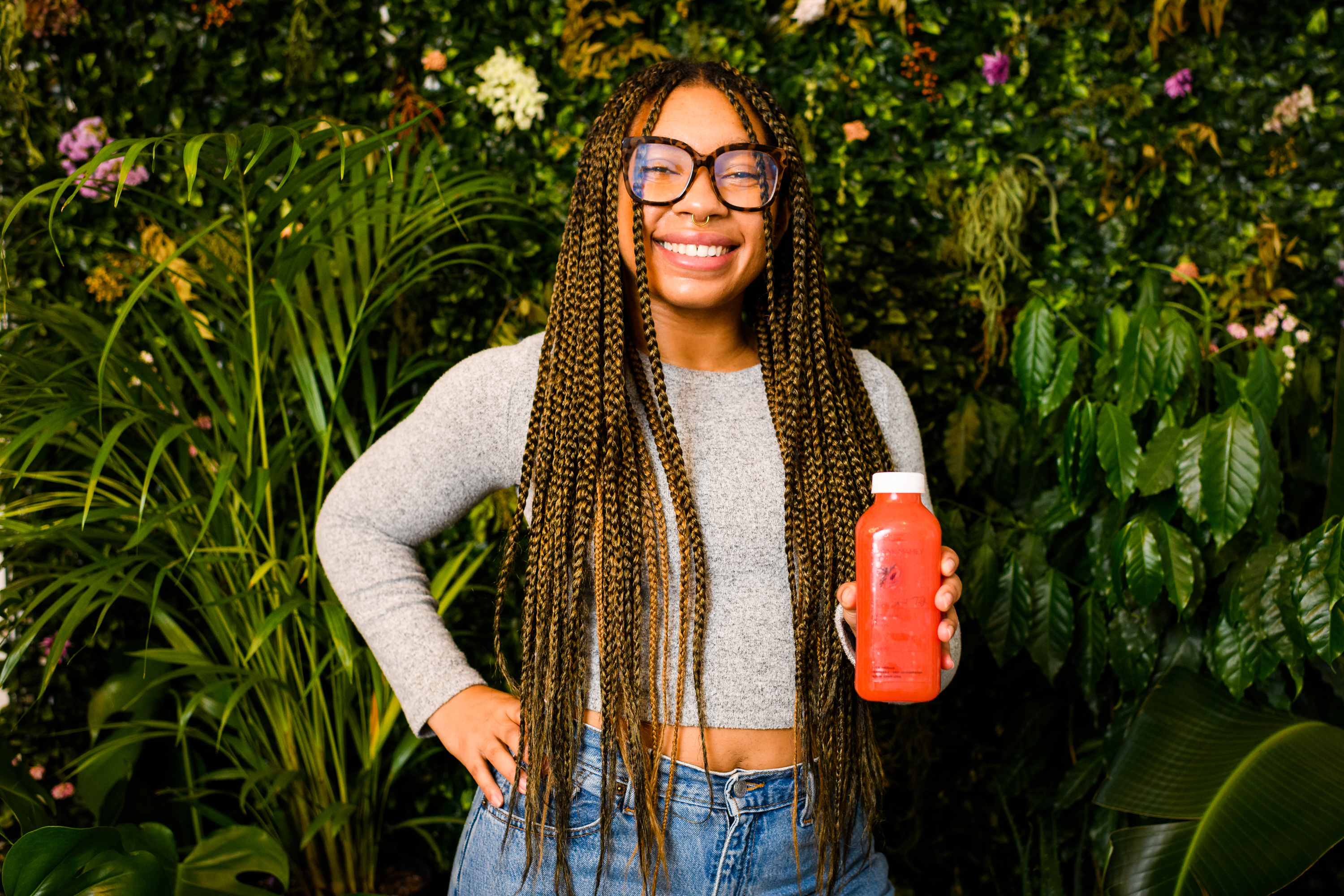 A woman with long dreads, glasses, and high-waisted jeans stands in front of a plant wall with a bottle of juice in her hand