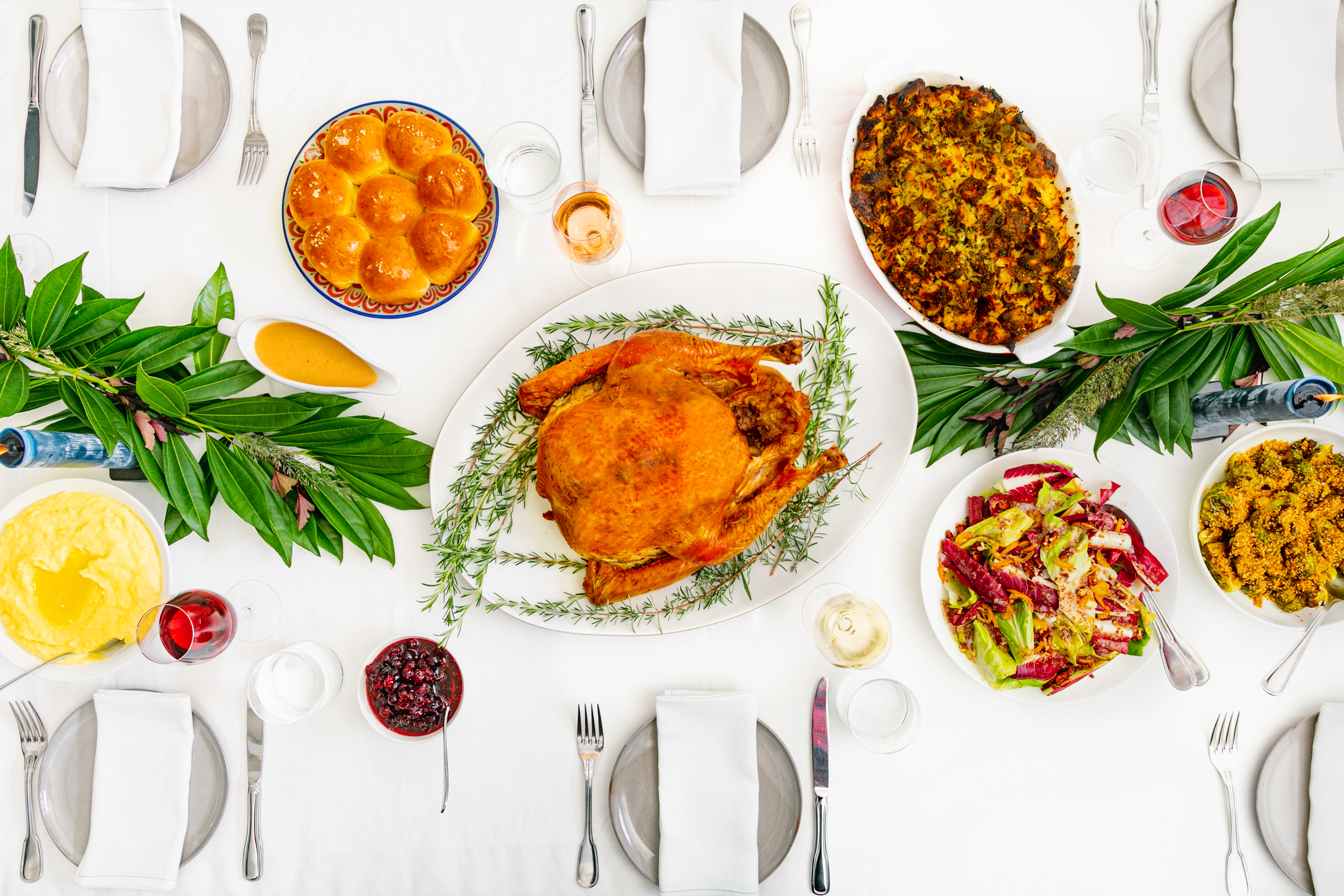 A Thanksgiving meal, including roasted turkey, bread rolls, salad, cranberries, and more, on a white tablecloth.