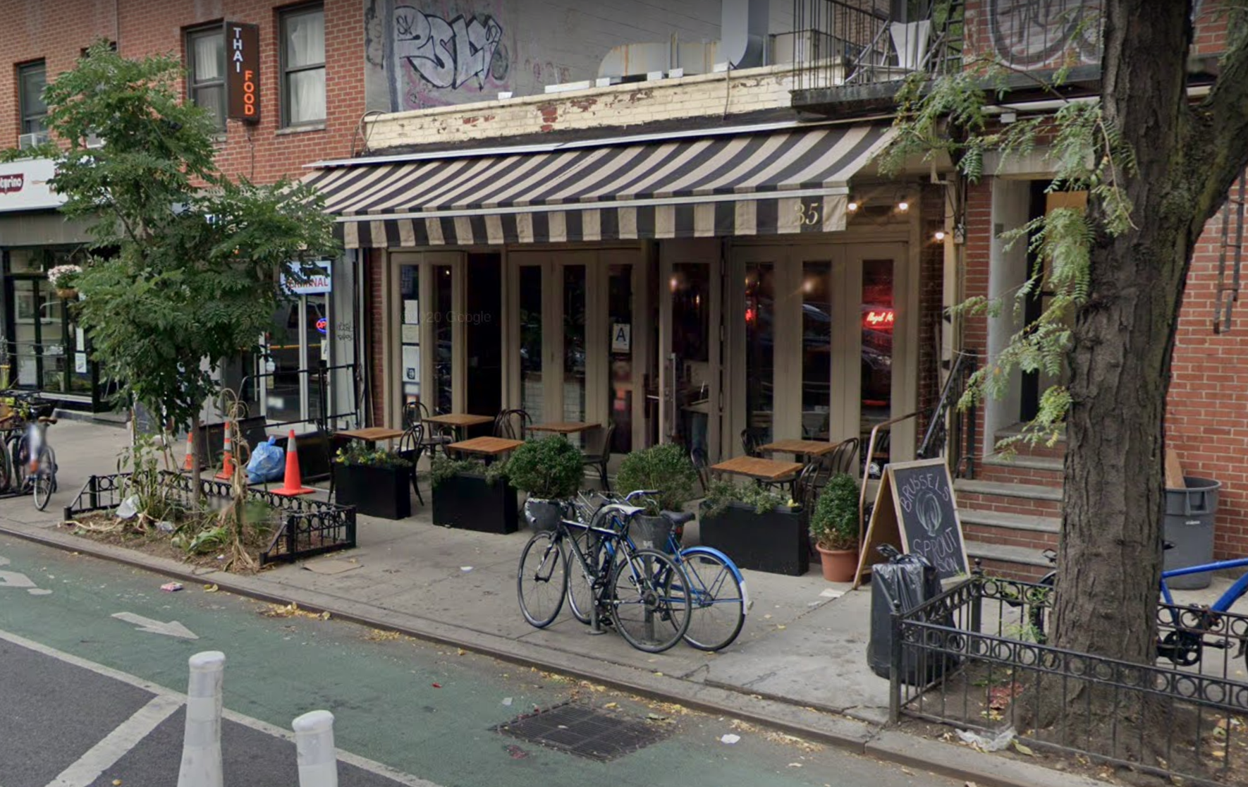 A bike lane runs along a sidewalk in front of a restaurant, where a striped black and white awning hangs over a small outdoor dining area