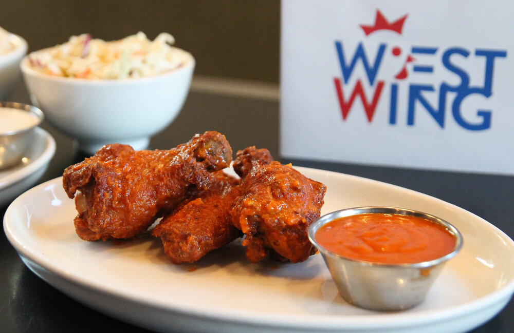 A plate of Buffalo wings with a side of sauce, mac salad, and a sign that says “West Wing” in the background