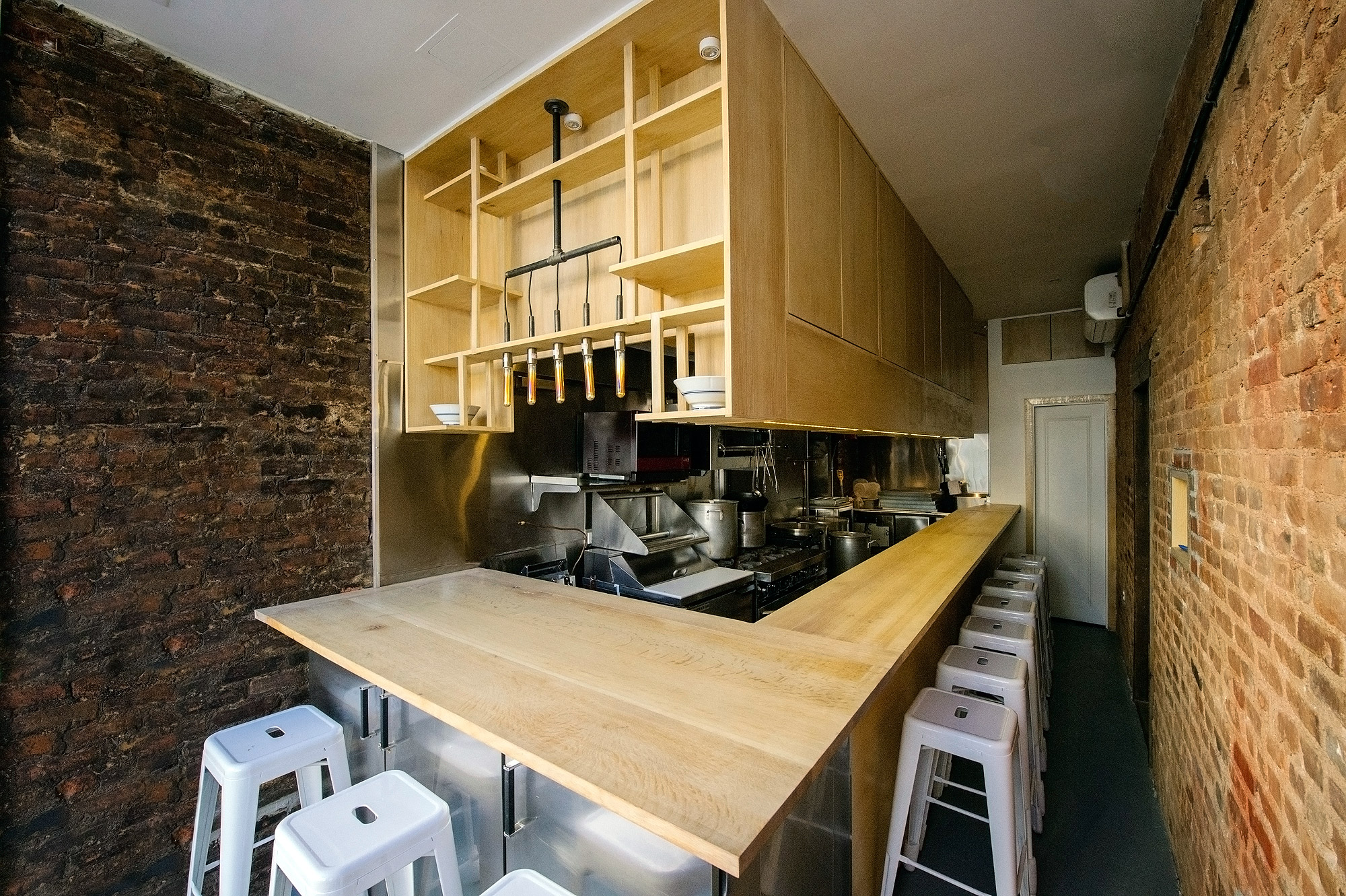 The interior of a snug ramen shop, with ten stools pulled up to a light wood counter. An open-air kitchen is visible in the background.