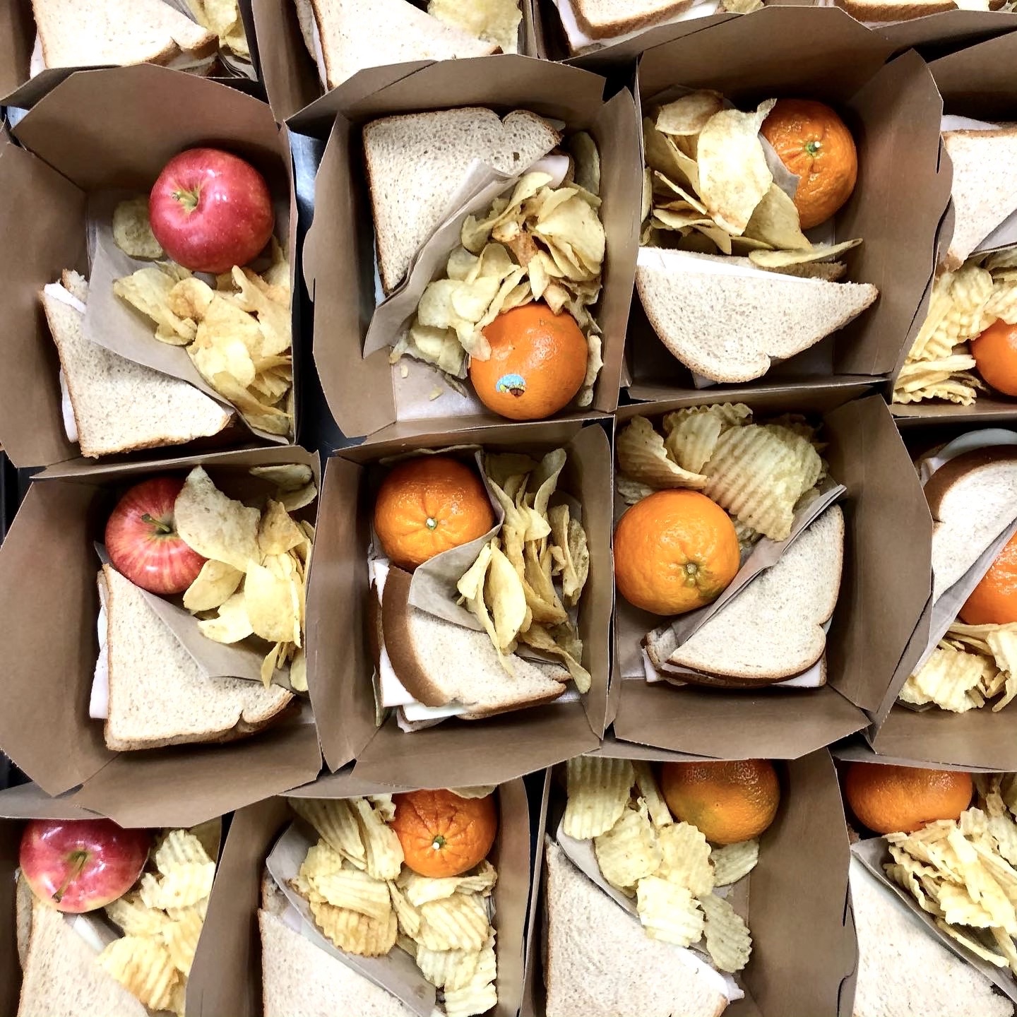 Brown takeout containers filled with crinkle cut chips, oranges, apples, and sandwiches sliced in half.