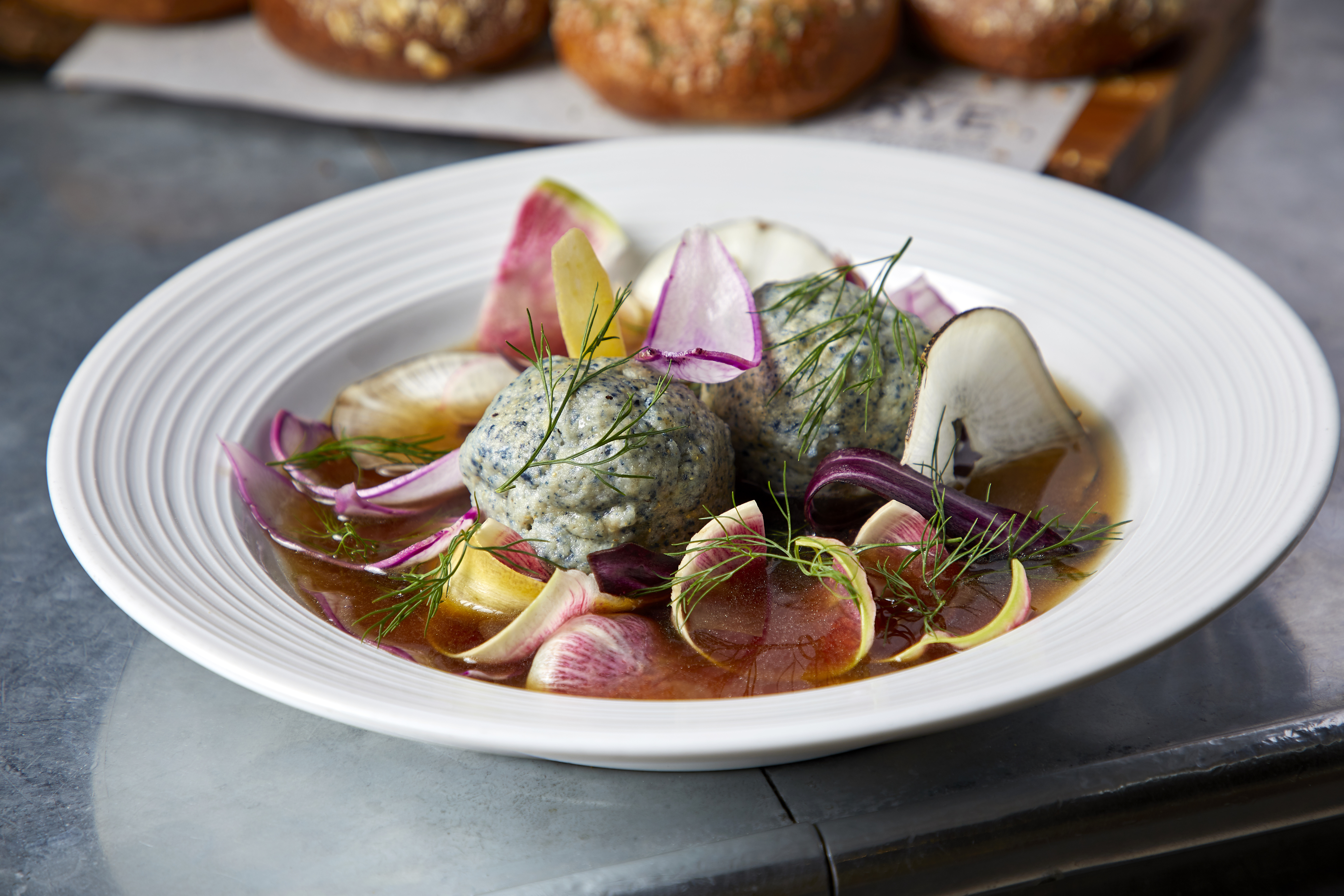 A white bowl of soup with blue matzo balls littered with thin-sliced vegetables.