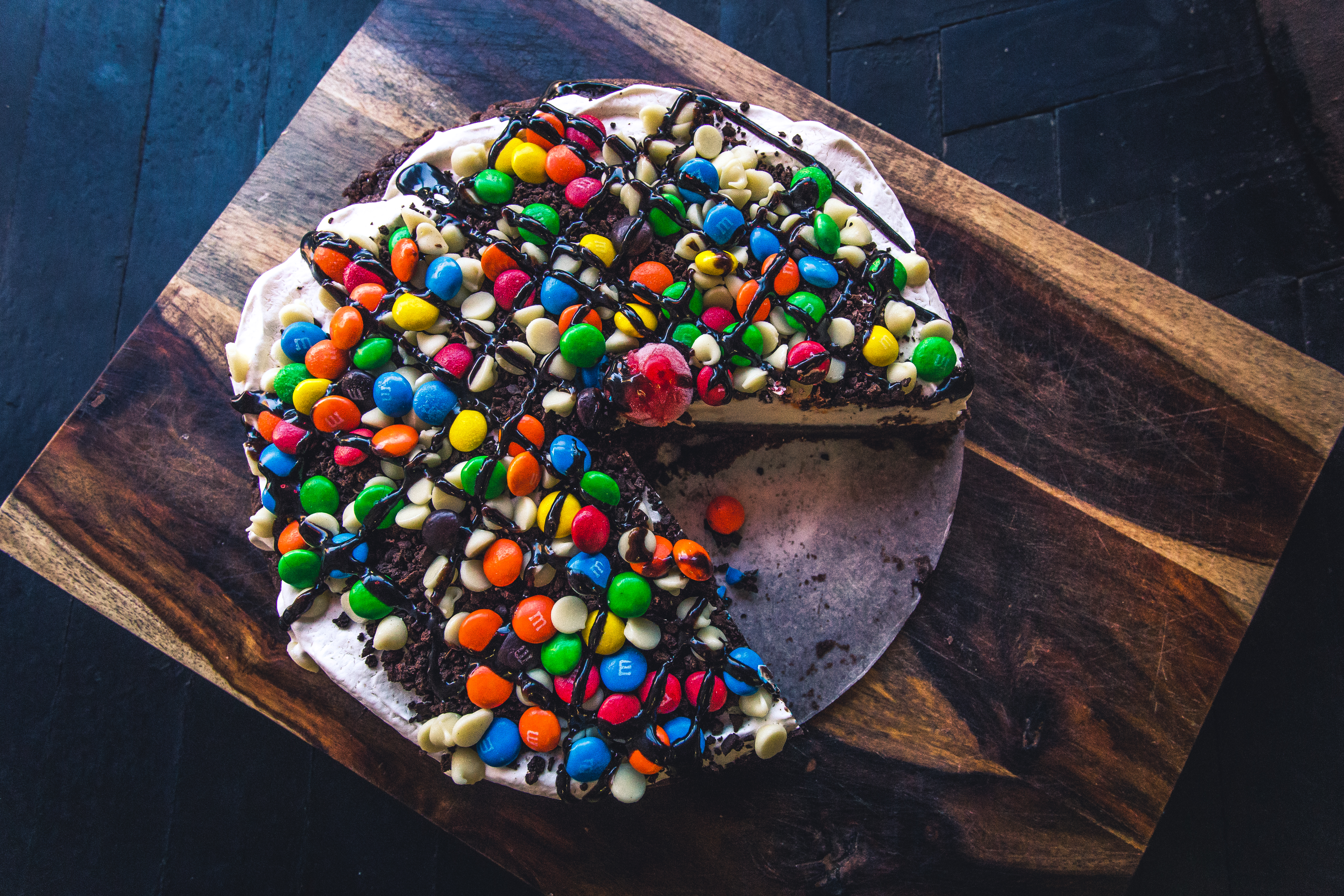 Overhead view of a round treat that combines a brownie crust with vanilla ice cream, candy, and chocolate drizzle. It’s on a wooden cutting board on top of a dark tabletop.
