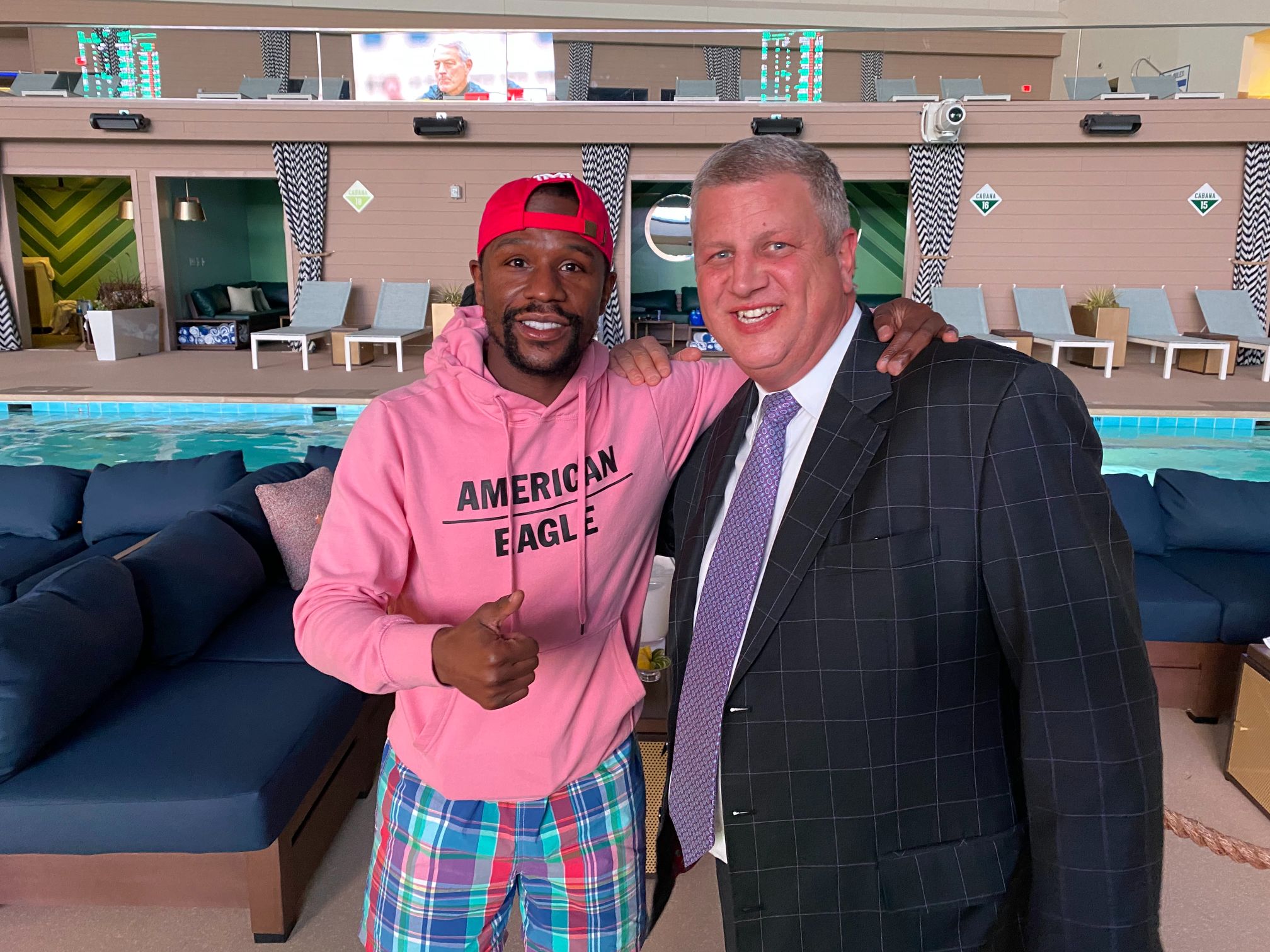 A man in a pink hoodie with a red ballcap and a man in a suit stand in front of a pool