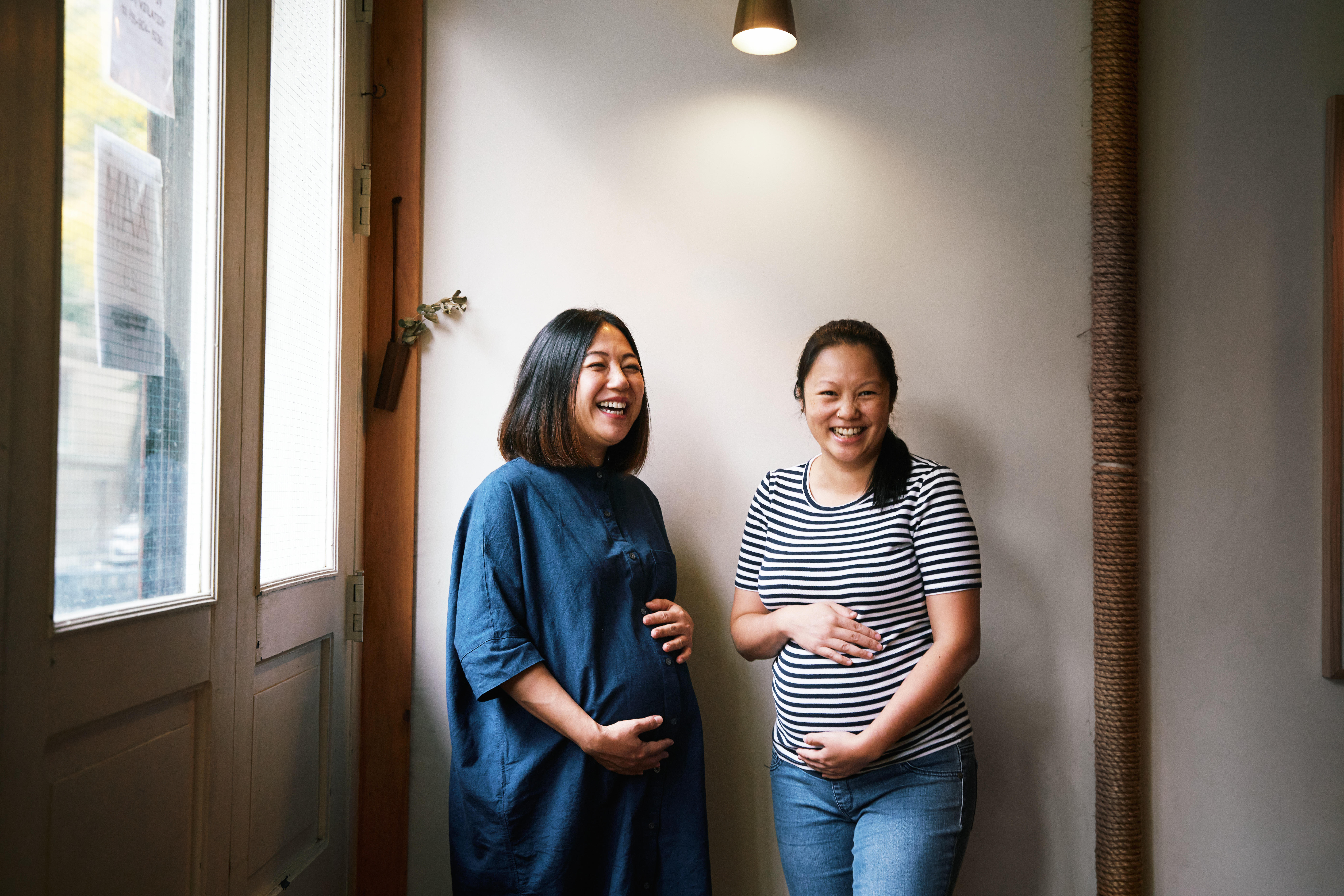 Two smiling and visibly pregnant women stand next to one another inside a restaurant