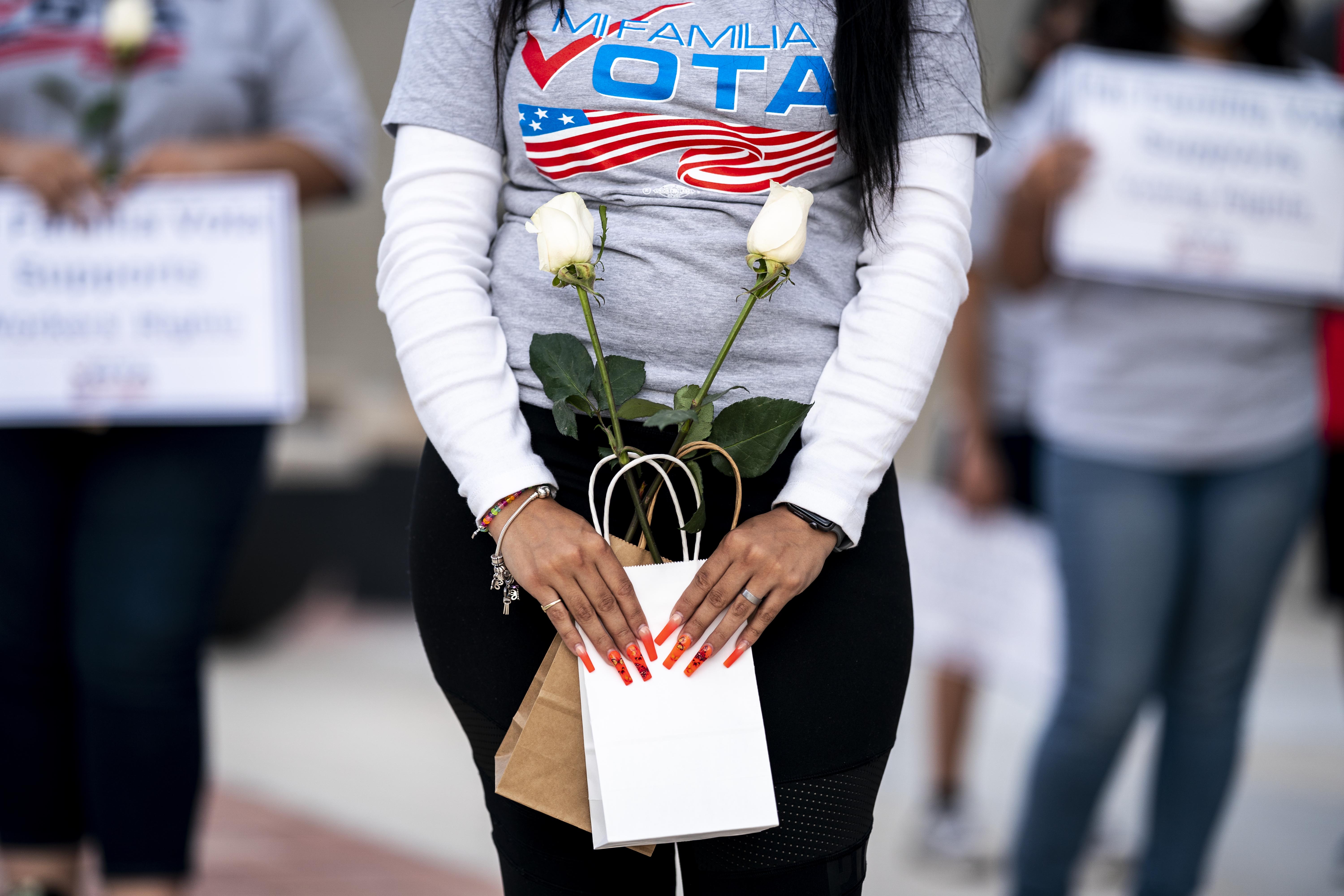 A Latina woman wearing a shirt that reads “Mi Familia Vota” and carrying two small bags with a white long-stem rose in each.