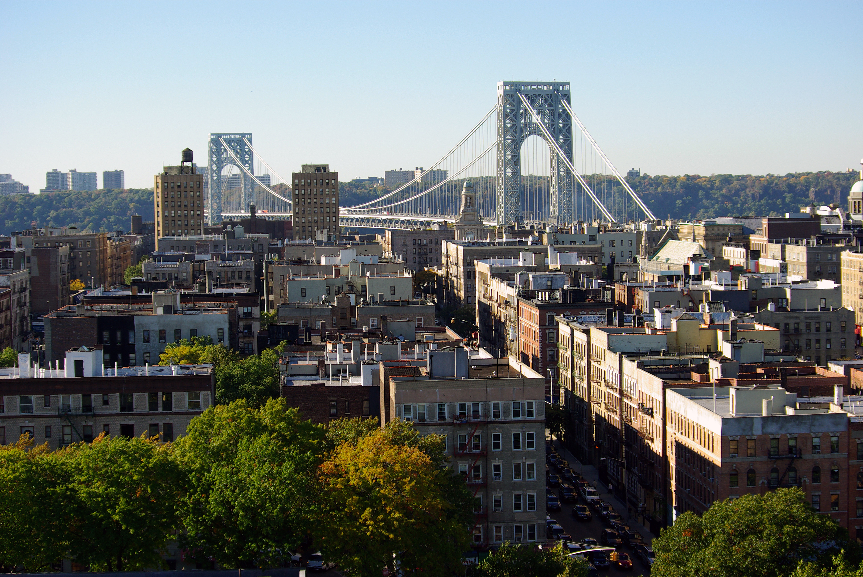 Several medium rise buildings and green trees are seen in the foreground with the George Washington Bridge in the background