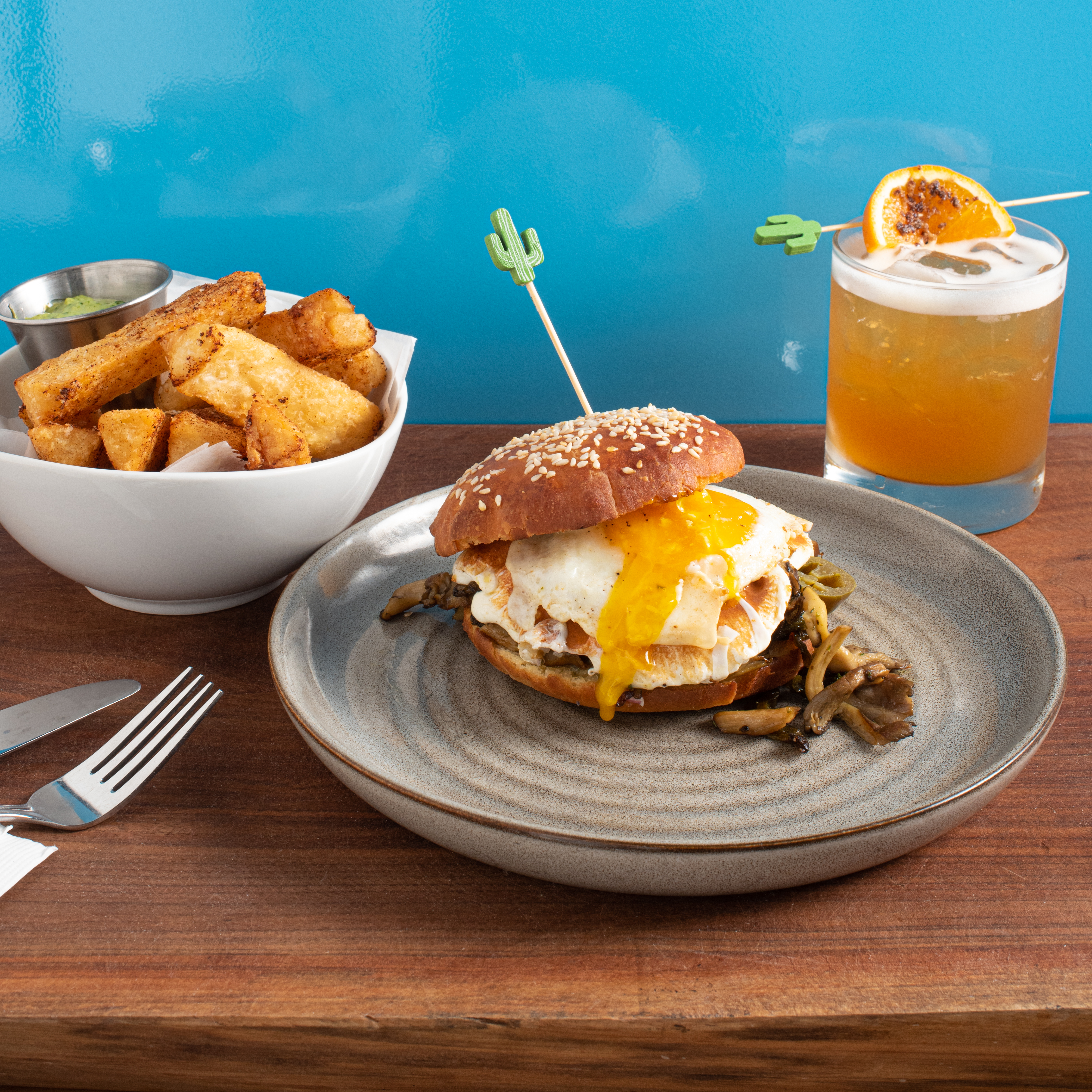 A breakfast sandwich with a gooey egg yolk on a bun is front and center, with an orange cocktail with a frothy top, and a white bowl of crispy potatoes in the background