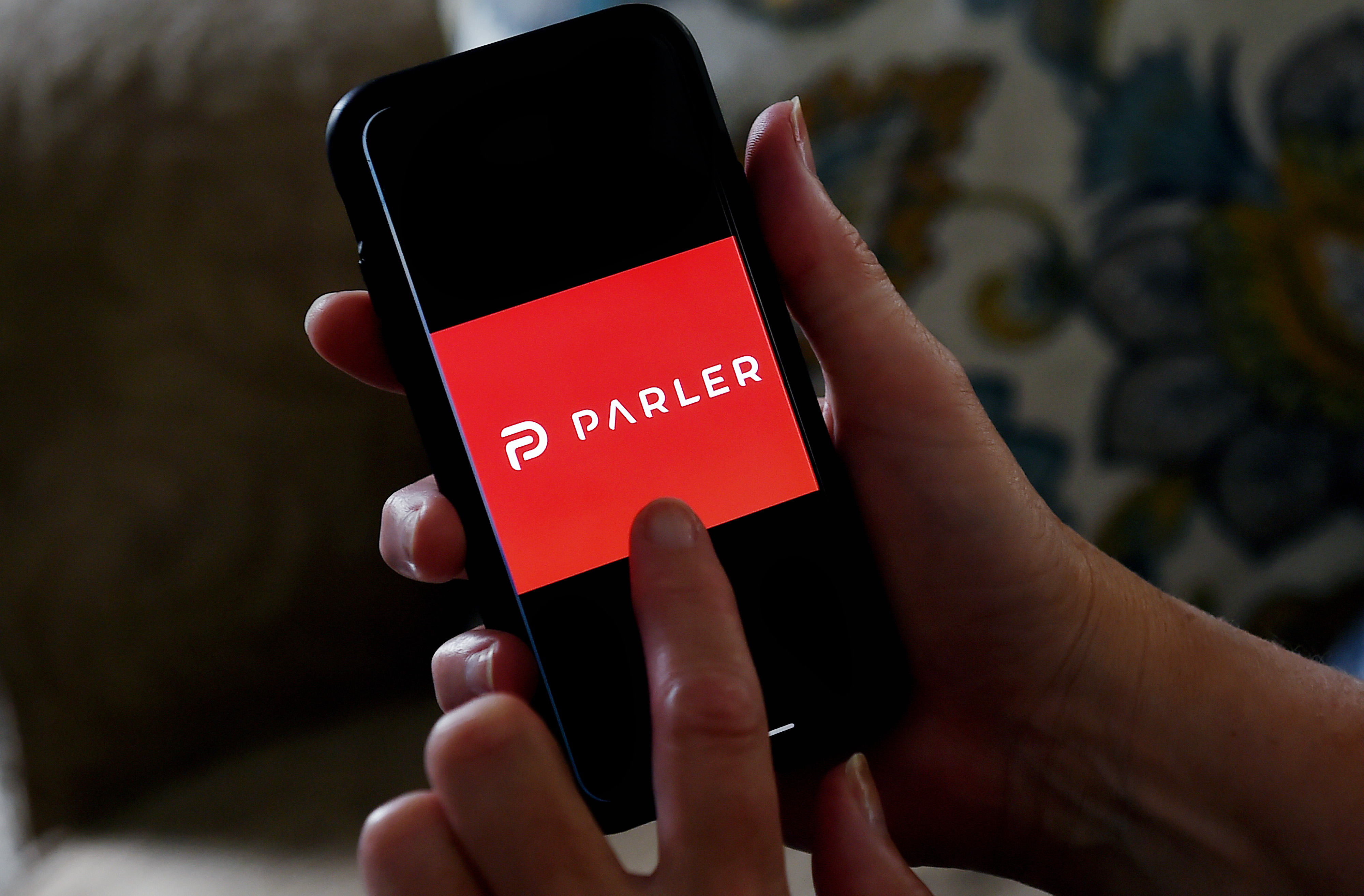 A person holds a smartphone and points to the logo for Parler on the screen.