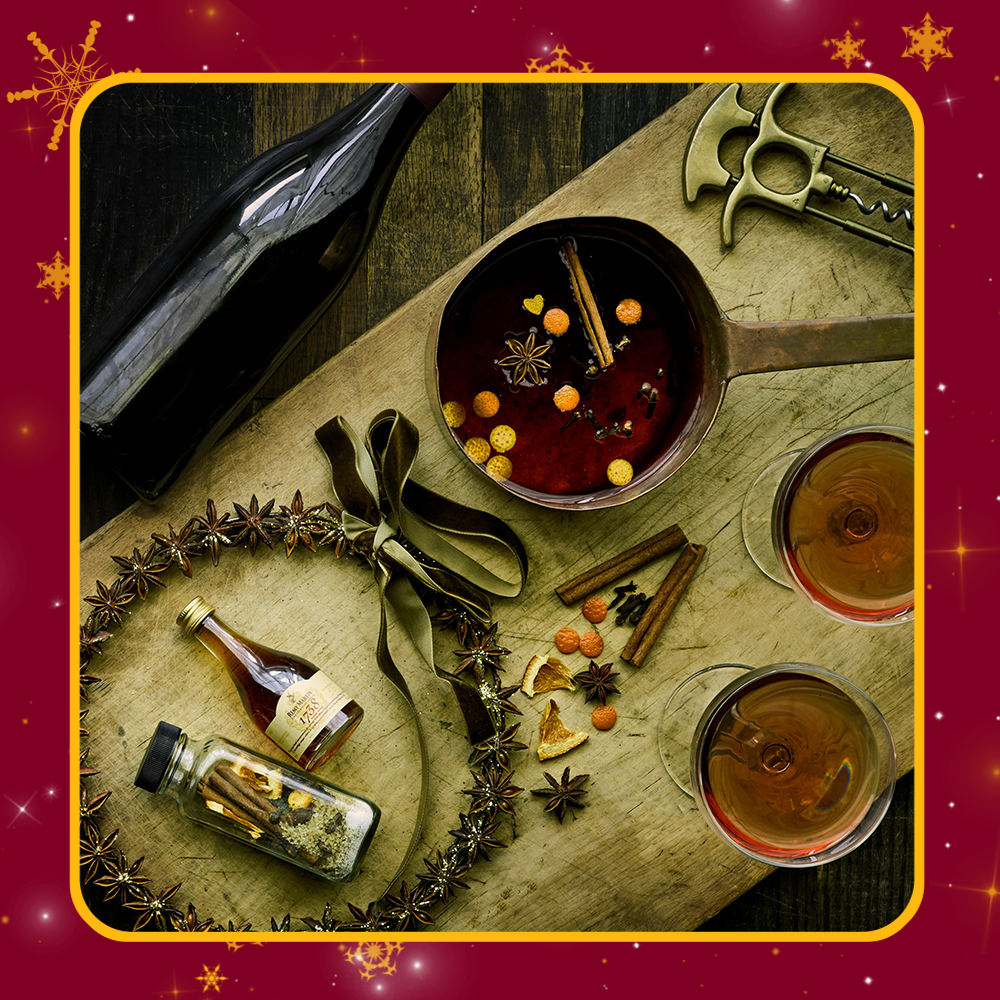 A photo of mulling spices in a pot with two coups filled with red liquid and containers of mulling spices and wine. The scene is very festive. 