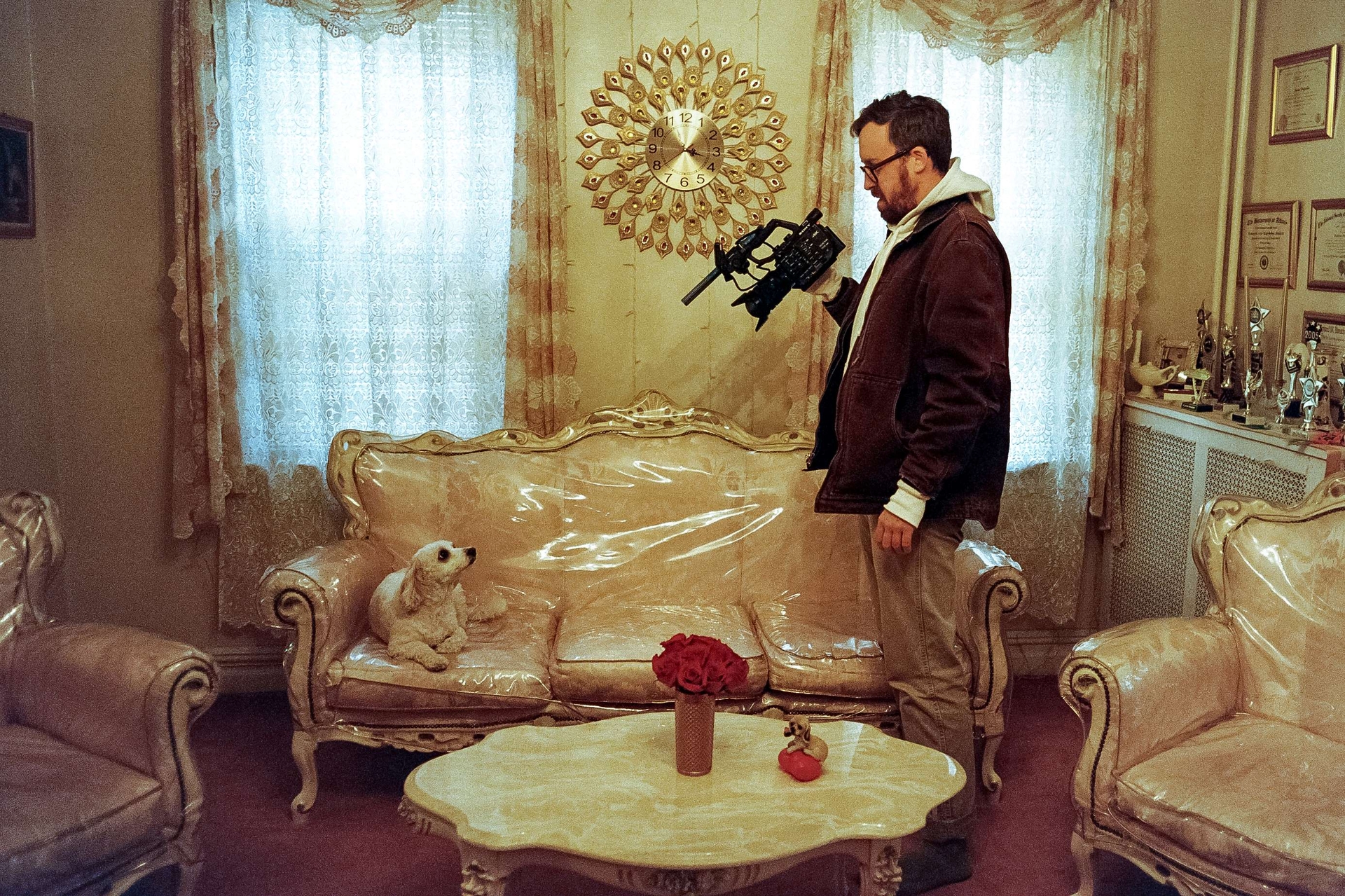 John Wilson stands with a camera filming a dog sitting on nice furniture covered with plastic wrap