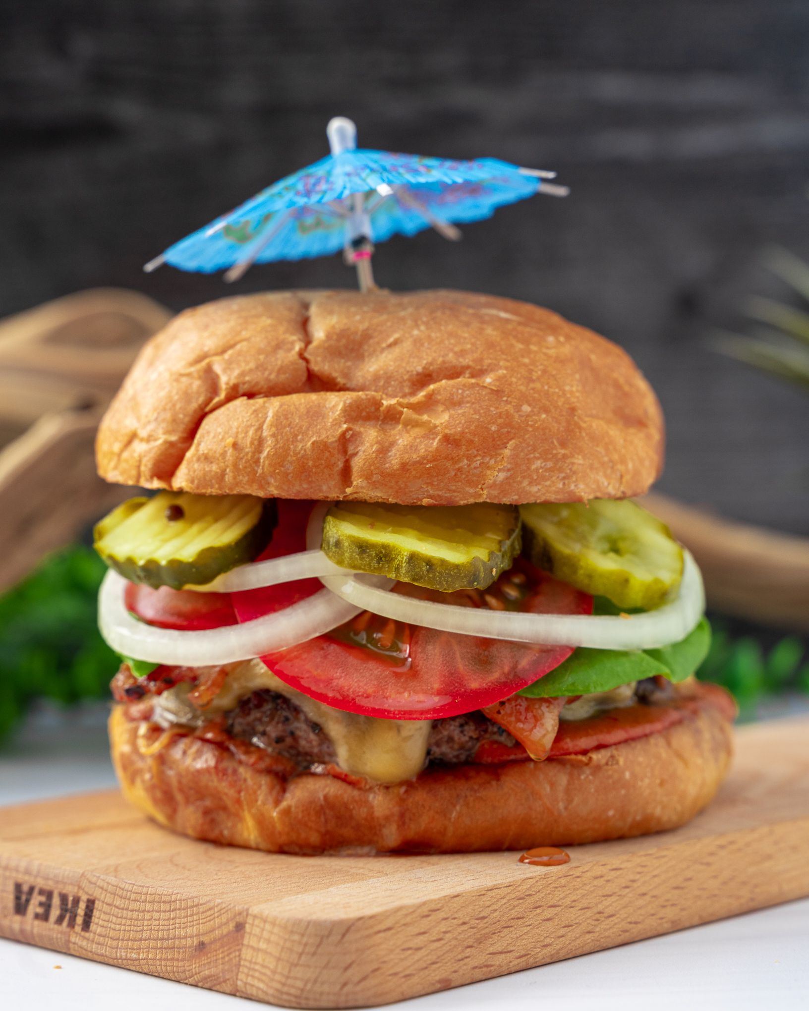 a burger with onions, pickles and tomatoes and topped with a blue cocktail umbrella