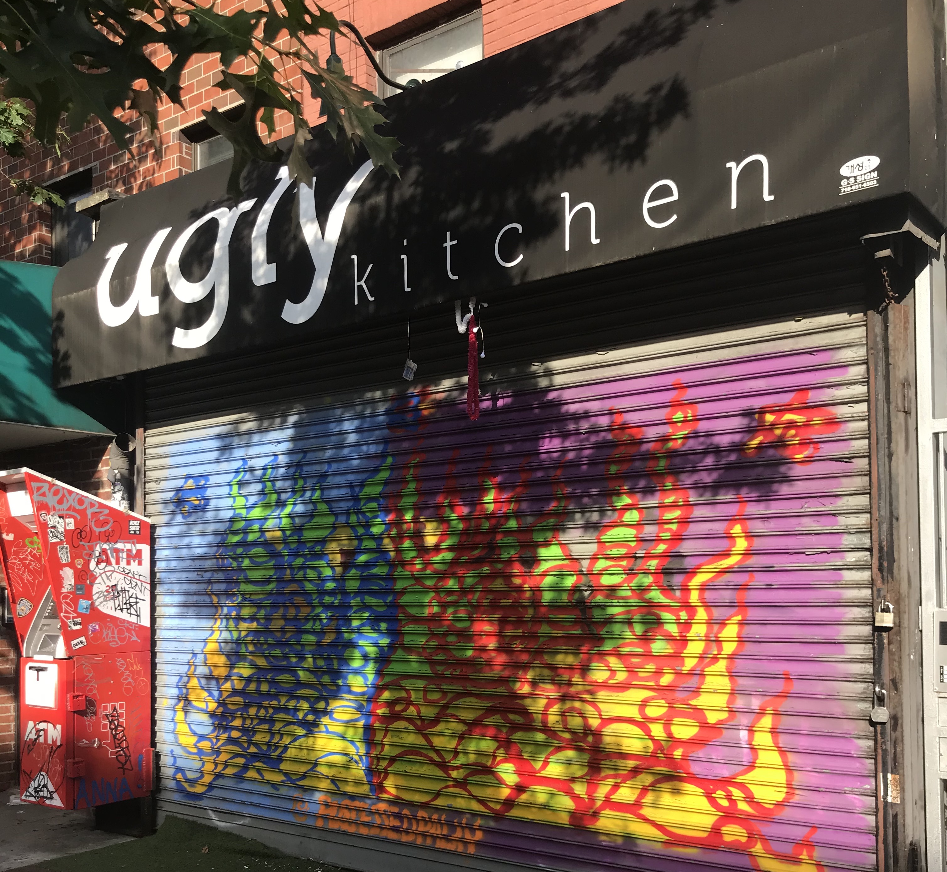 The exterior of a restaurant with a black awning and white lettering reading Ugly Kitchen. The front gate is pulled down with colorful graffiti in view on the gate. There is a red ATM to the left of the gate.