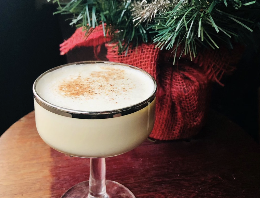 A glass of eggnog sprinkled with cinnamon and a Christmas wreath and red bow in the background