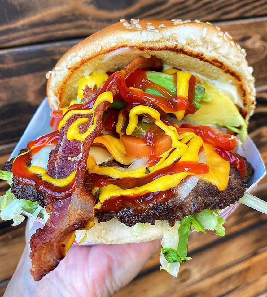 A giant burger stuffed with bacon, avocado, peppers, onion, shredded lettuce, with ketchup and mustard on a sesame seed bun. The burger is so stuffed with toppings it looks like it’s in danger of everything spilled out of it