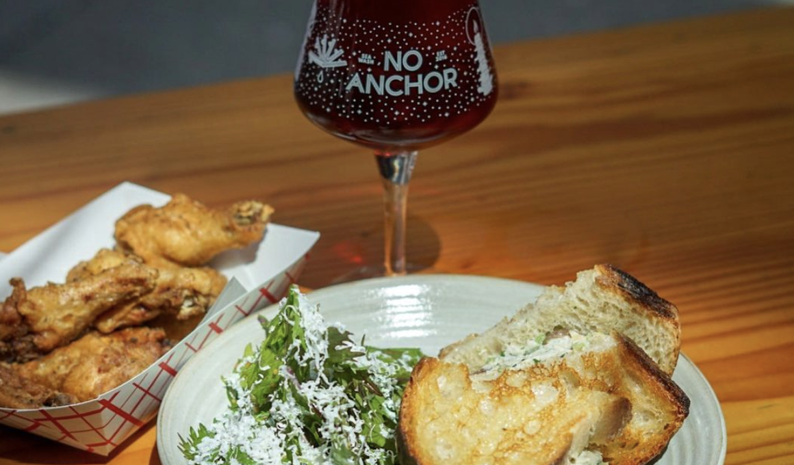A glass of beer that says “No Anchor” next to a paper tray filled with wings and a sandwich on a plate with a side salad