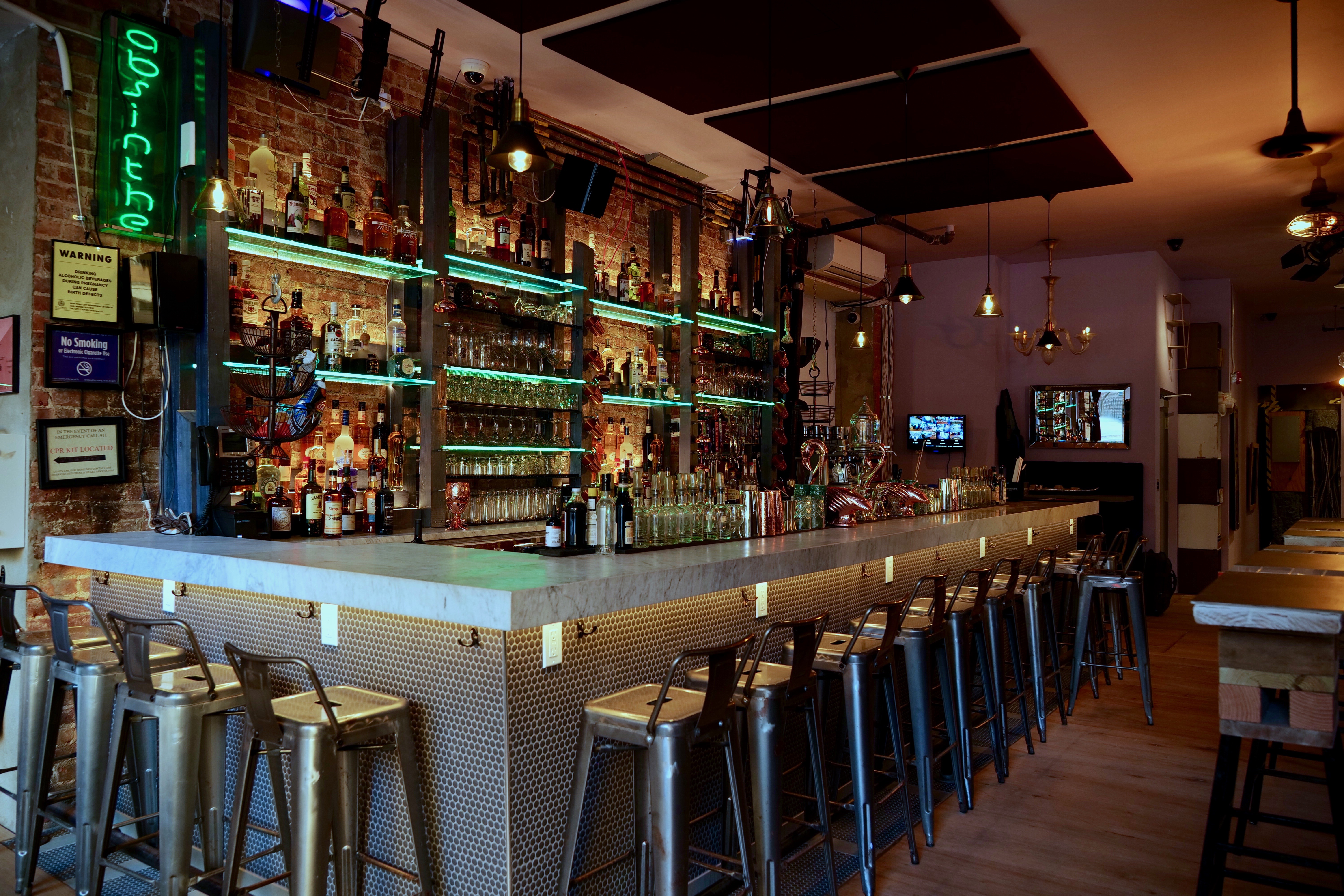 An interior restaurant photo focusing on a backlit bar with alcohol lining the shelves and backless stools set up around the outer perimeter of the bar