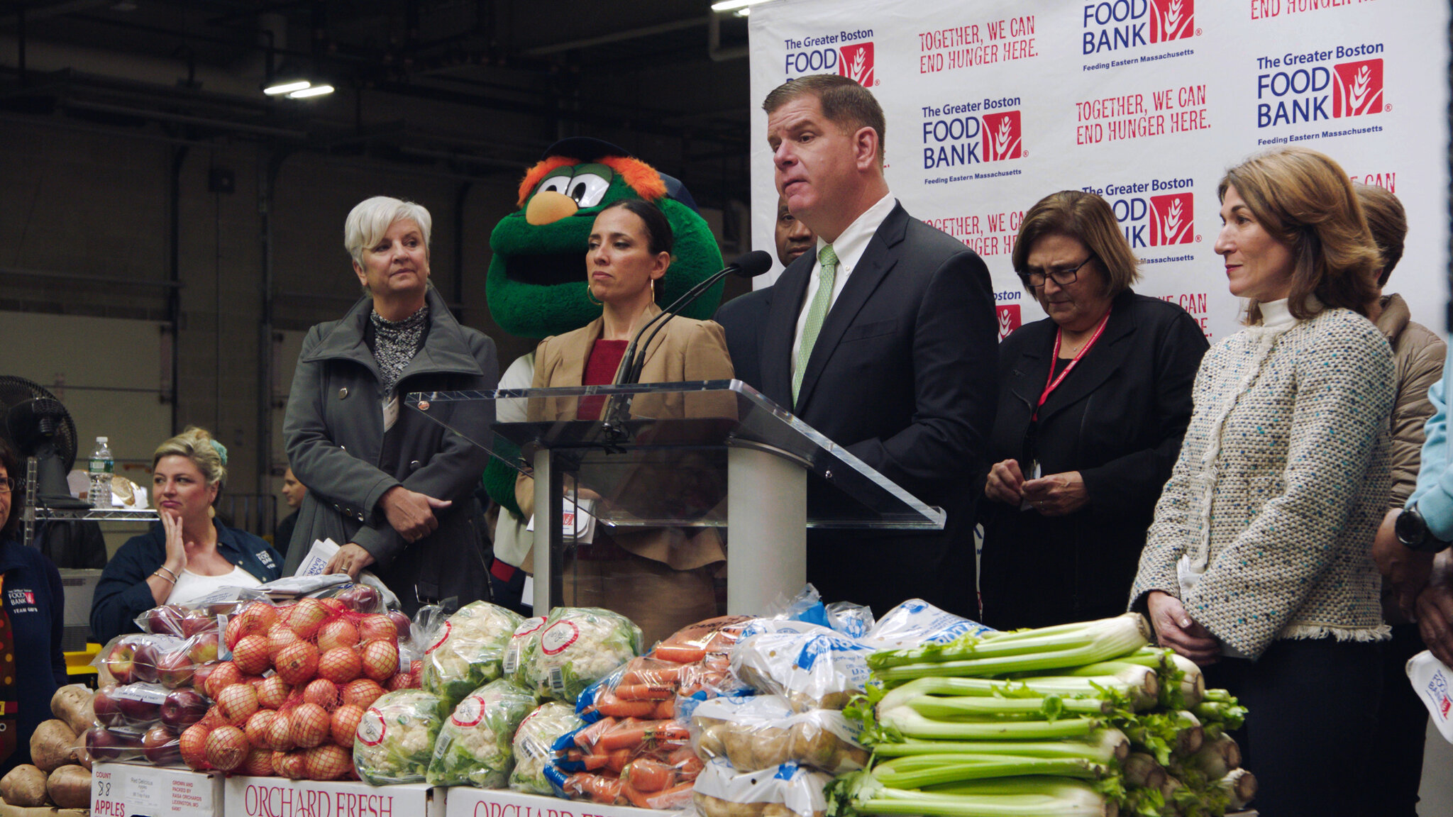 The mayor stands at a podium with several associates, flanked by the Green Monster mascot and piles of vegetables.