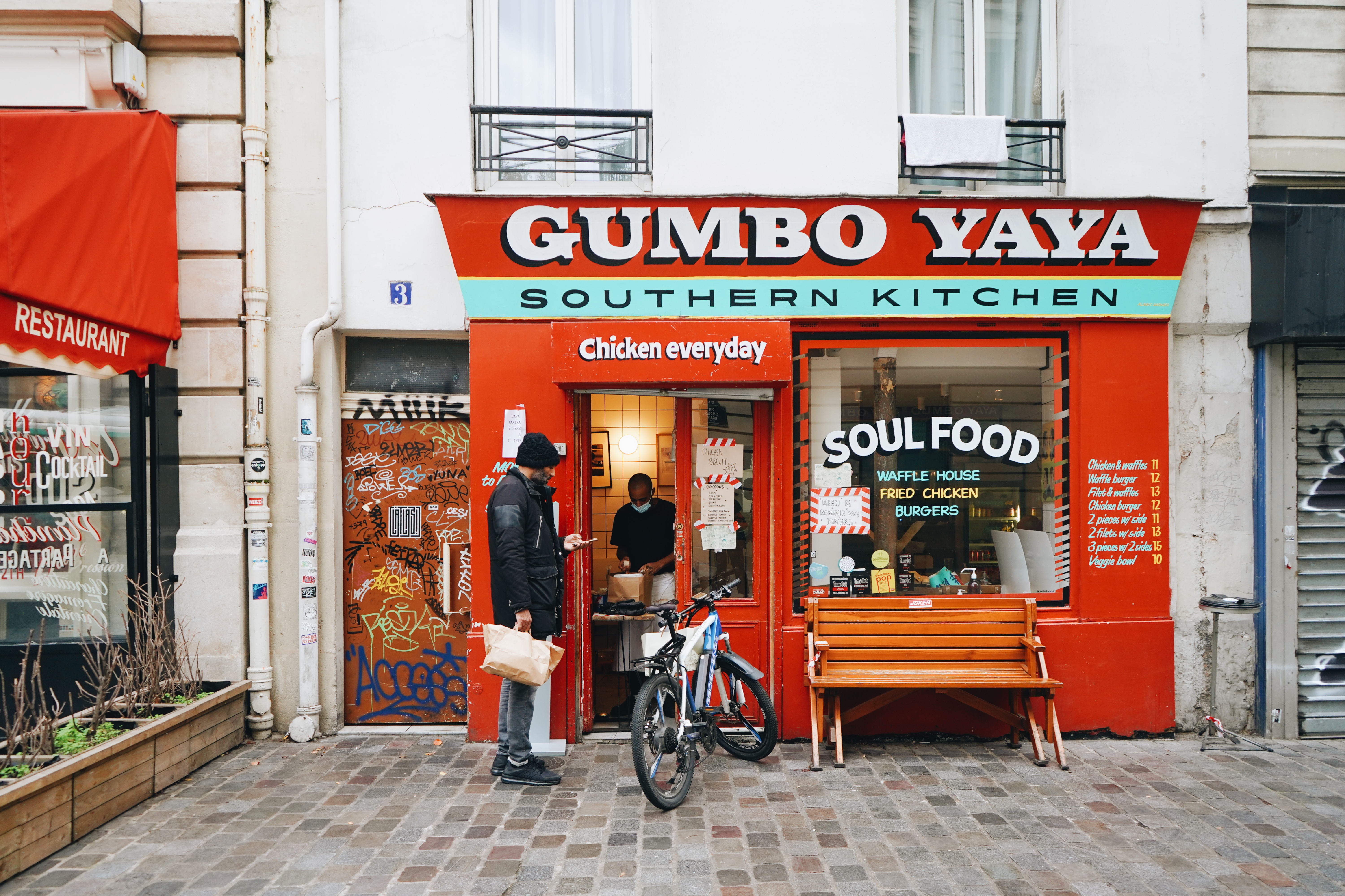 A man stands next to a bicycle in the brick plaza outside of Gumbo Yaya as another man inside the doorway packs up some food. The restaurant’s bright-red facade says “Gumbo Yaya Southern Kitchen” and its front window reads “Soul Food.”