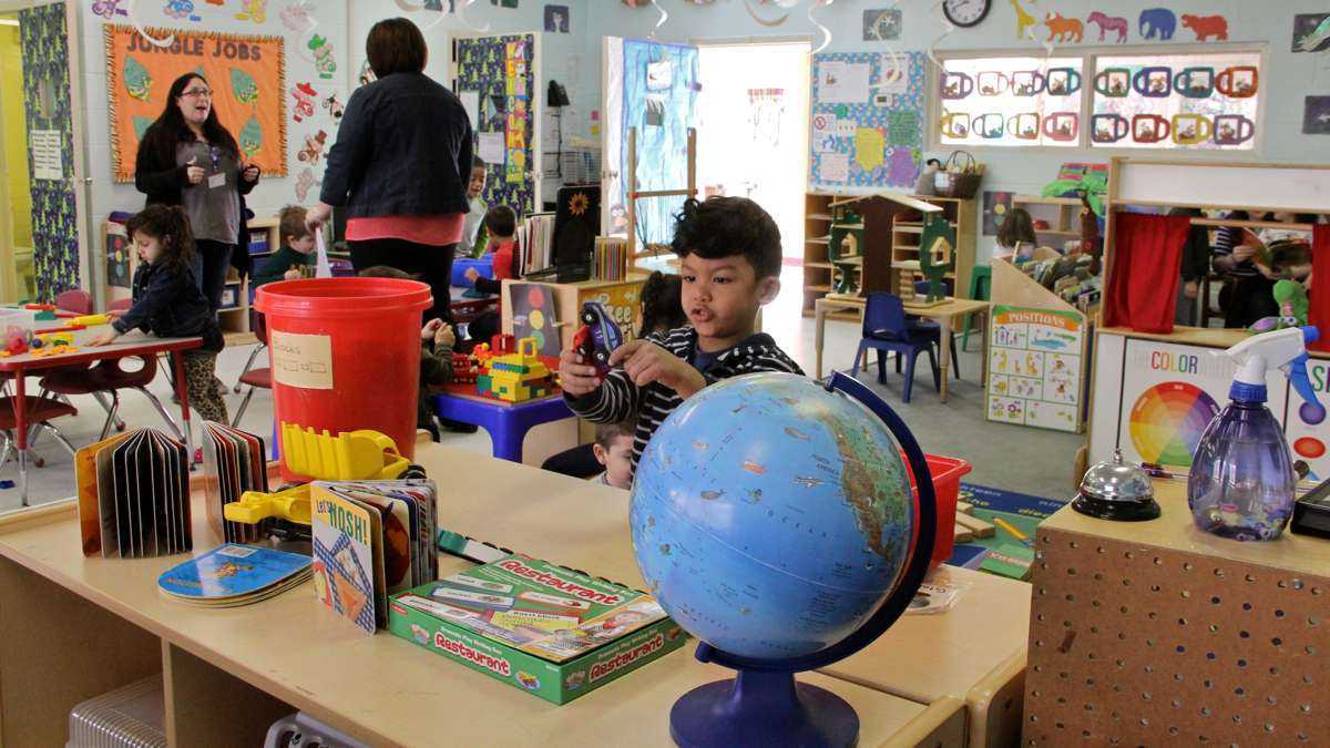 A boy plays in a classroom, just behind a large globe. There are two adults speaking and other kids playing in the classroom.