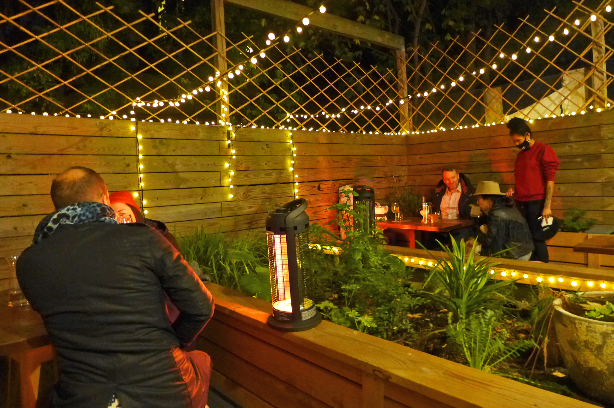 A darkened and fenced backyard strung with lights and with two tables of diners.