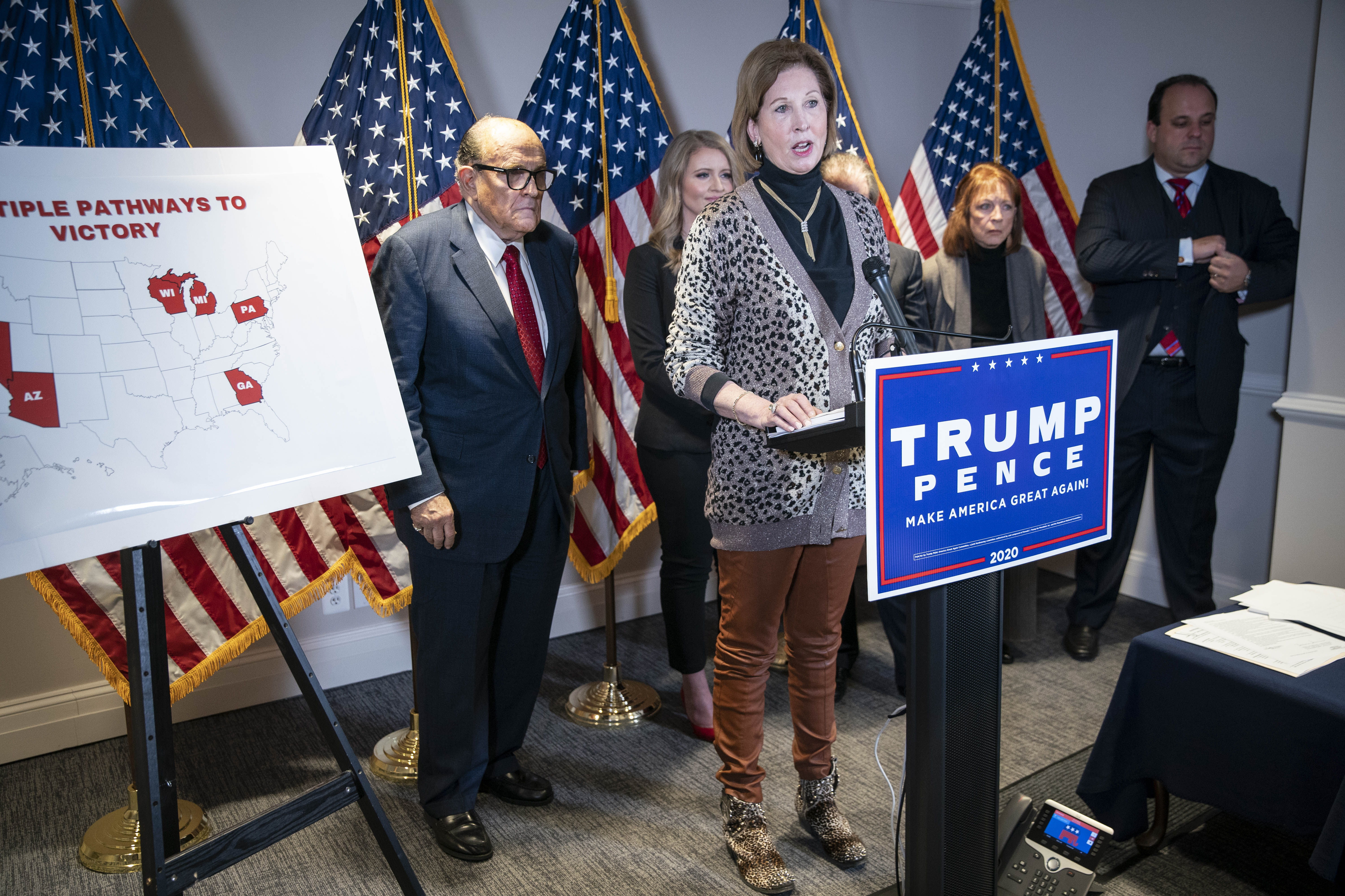Powell, in a leopard print sweater, black turtleneck, and tan slacks, speaks at a podium with a Trump Pence sign. Behind her are a row of US flags and Rudy Giuliani in a dark suit and red tie.
