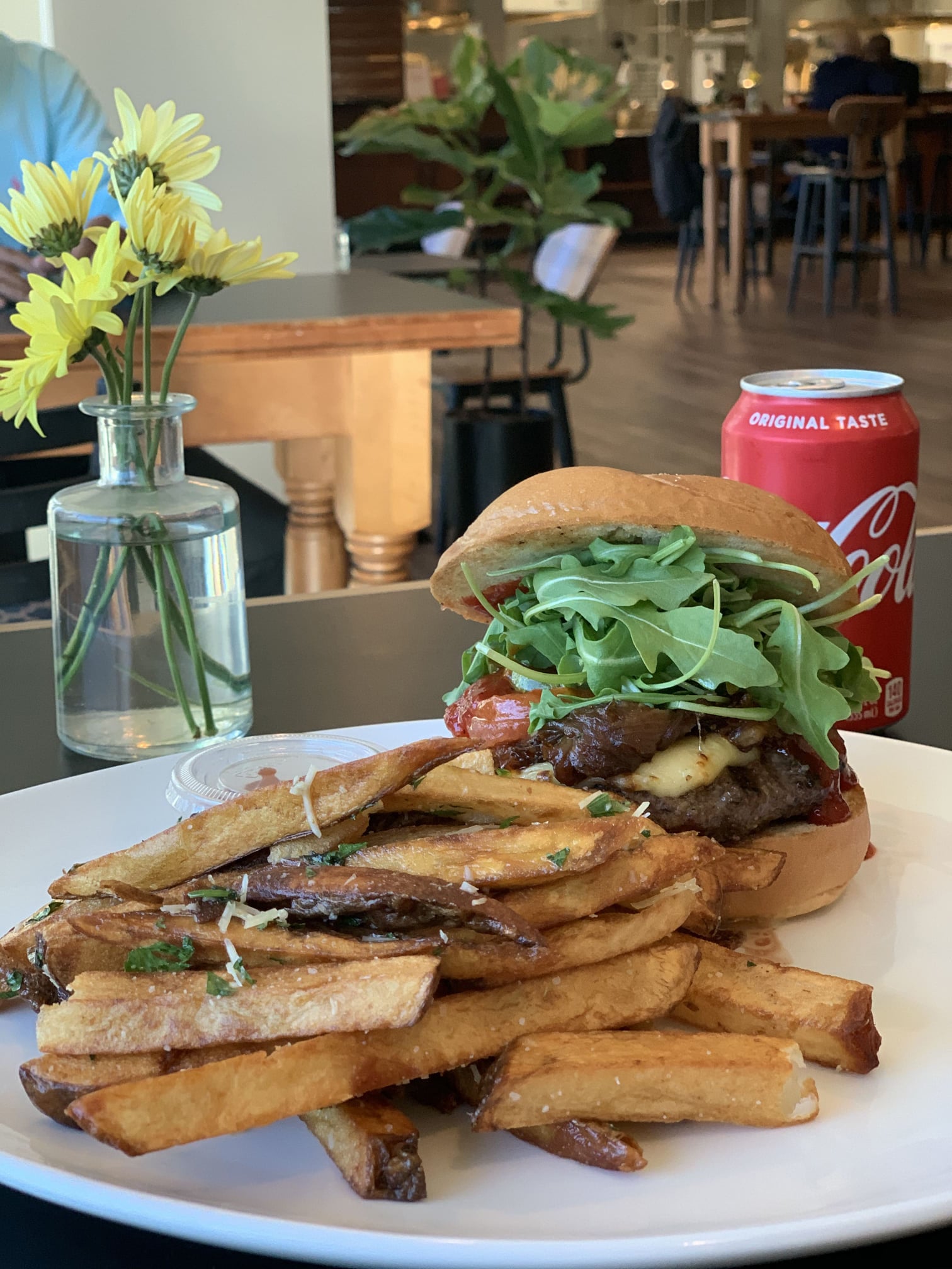 A double stack burger topped with leaf green arugula, smoked bacon, and cheese served with a side of steak fries. A red Coca Cola can sides beside the burger and fries with a clear vase with yellow daisies
