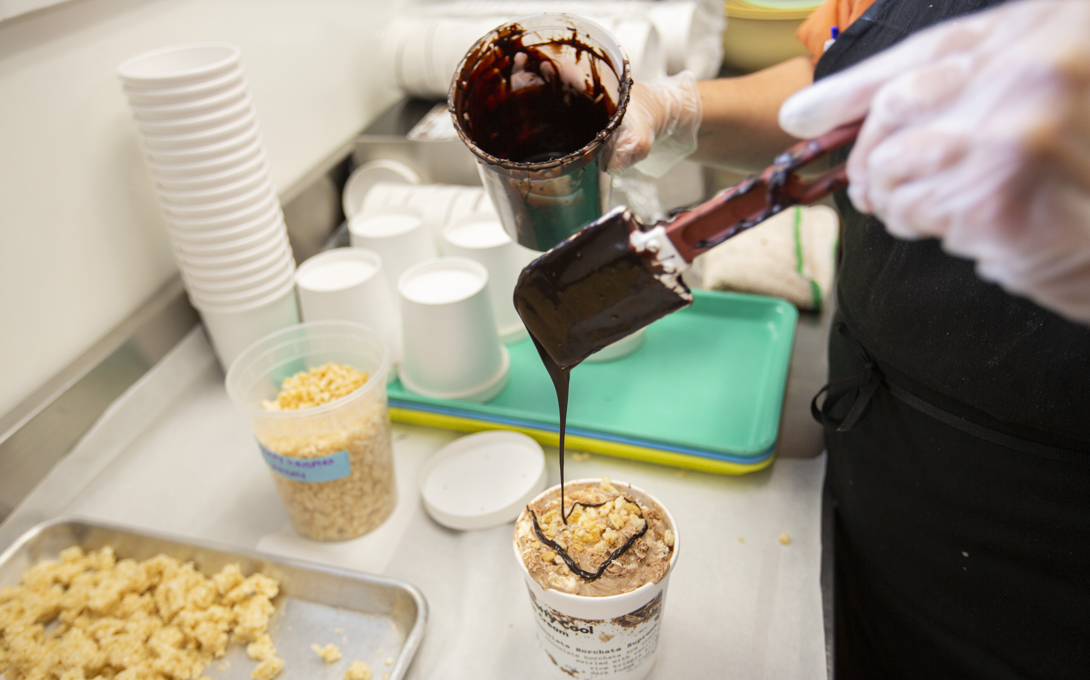 A person wearing latex gloves drizzles chocolate over a pint of ice cream in a white container.